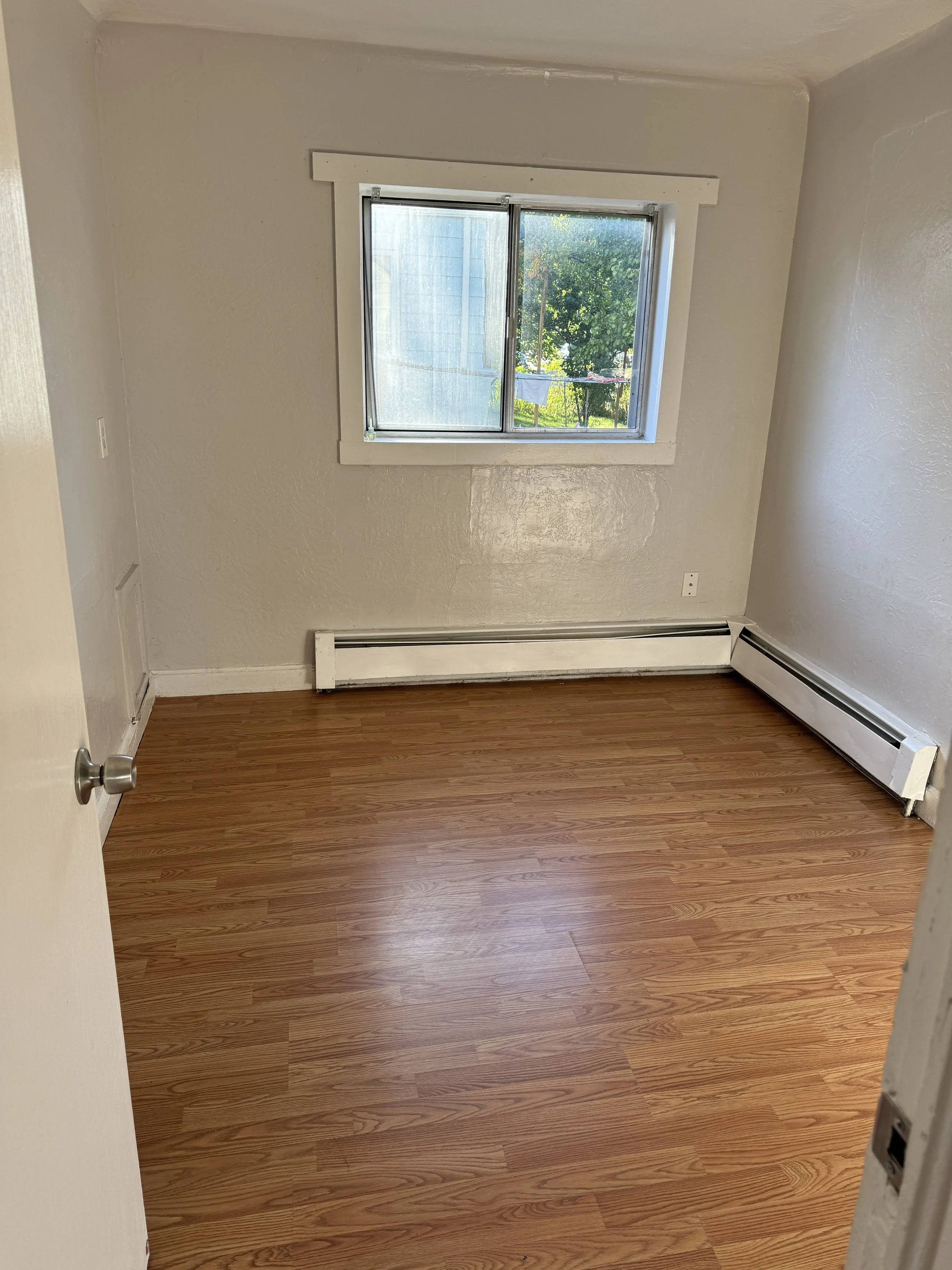 Empty room with hardwood floors, a window with white trim, and a baseboard heater along the wall.