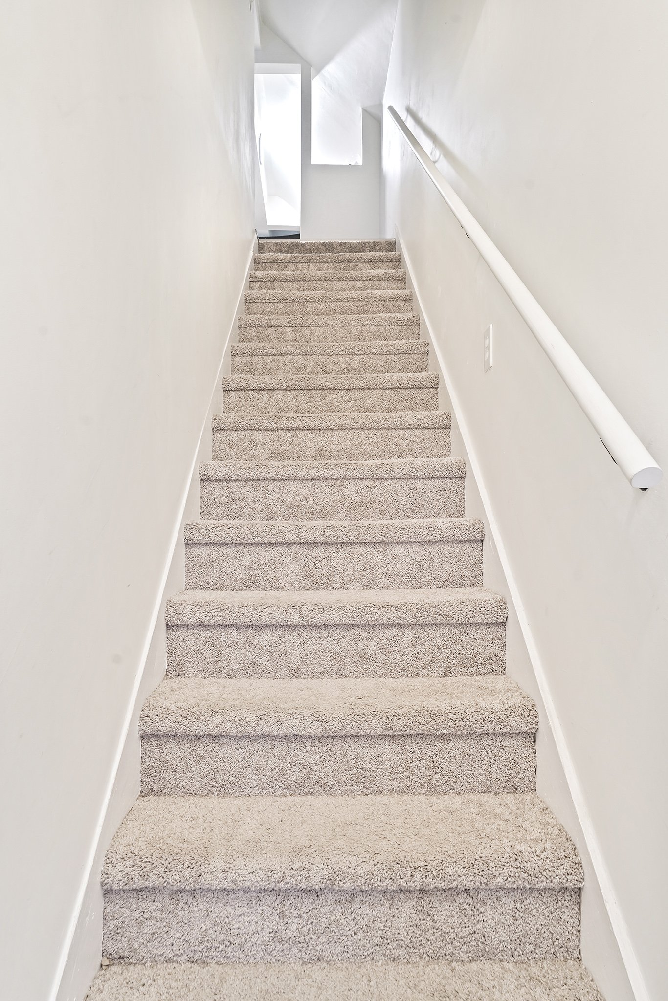 Indoor staircase with gray carpet, white walls, and a white handrail on the right side. A window at the top lets in natural light.