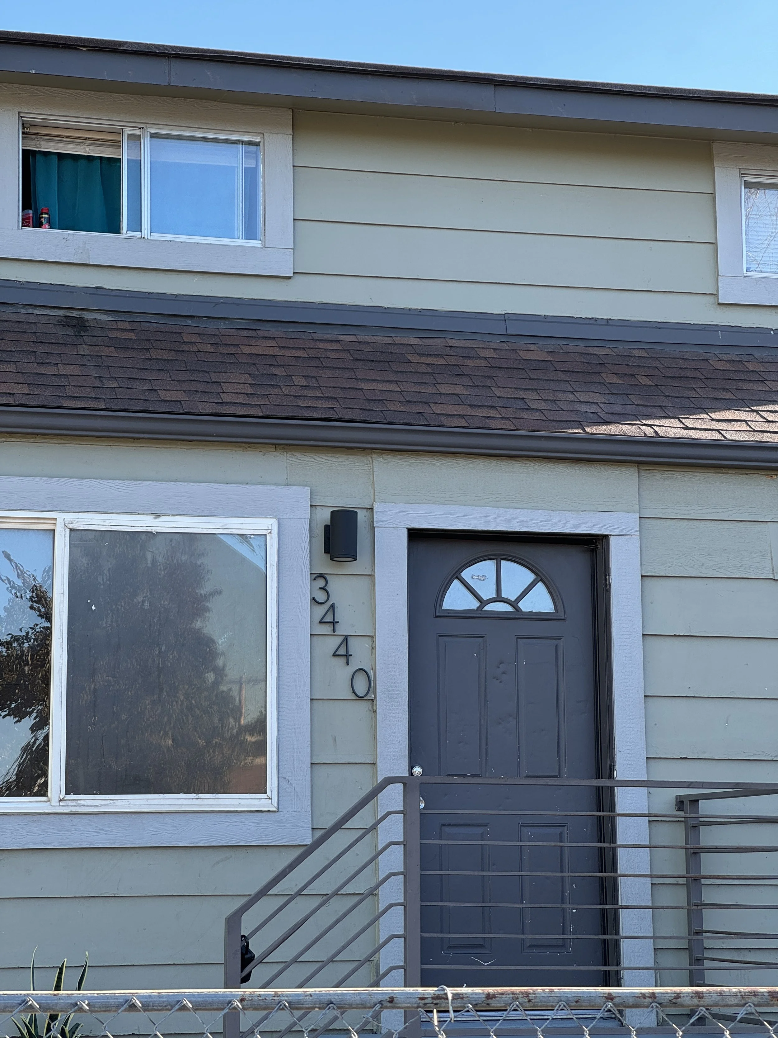 Front view of a two-story house with beige siding, a window with blue curtains, and a black front door with house number 3440. There is a railing in front and a small porch.