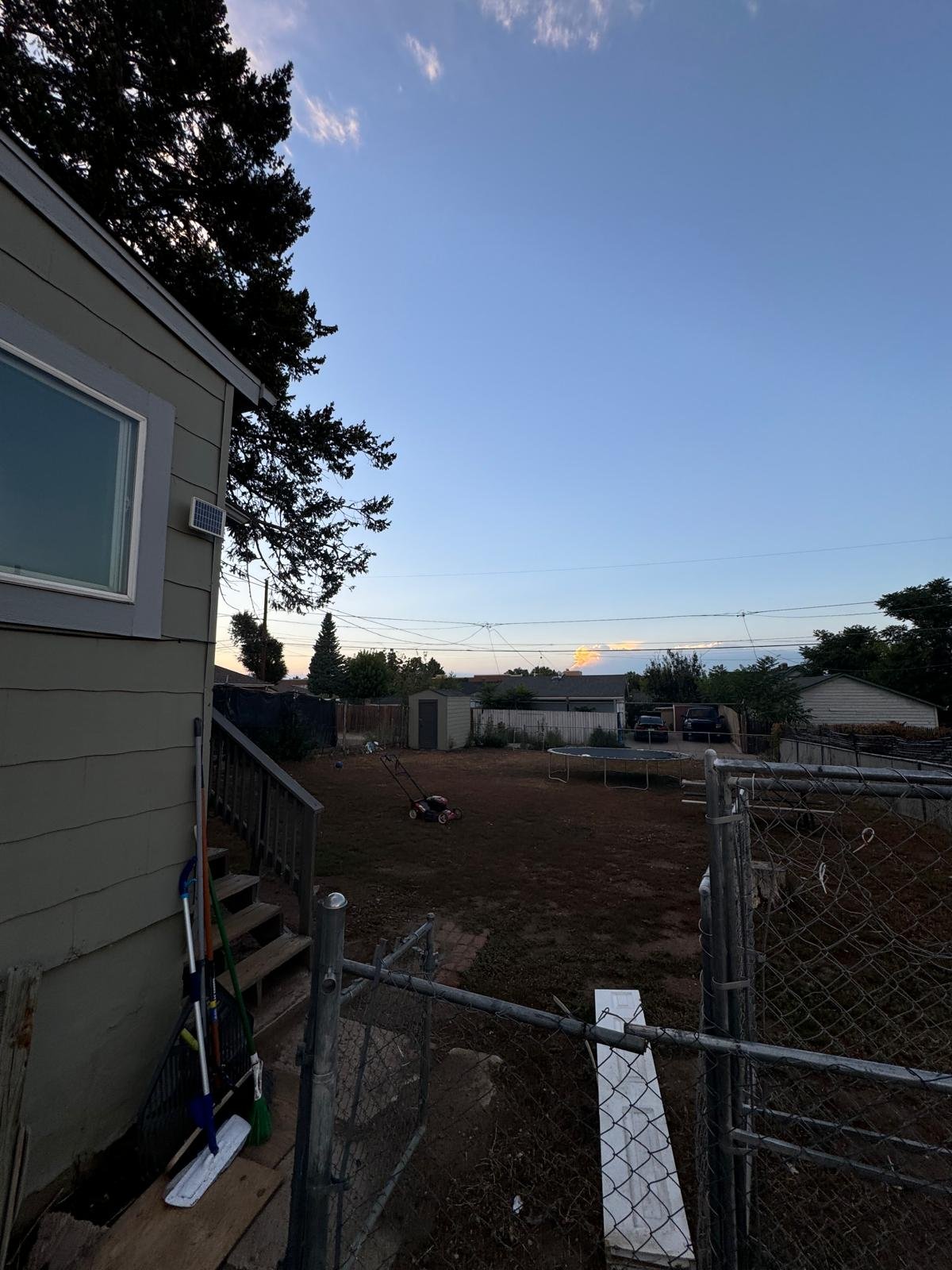 A backyard with a muddy brown lawn, a trampoline, a small shed, and a few trees and shrubs. The sky is clear with some clouds on the horizon.