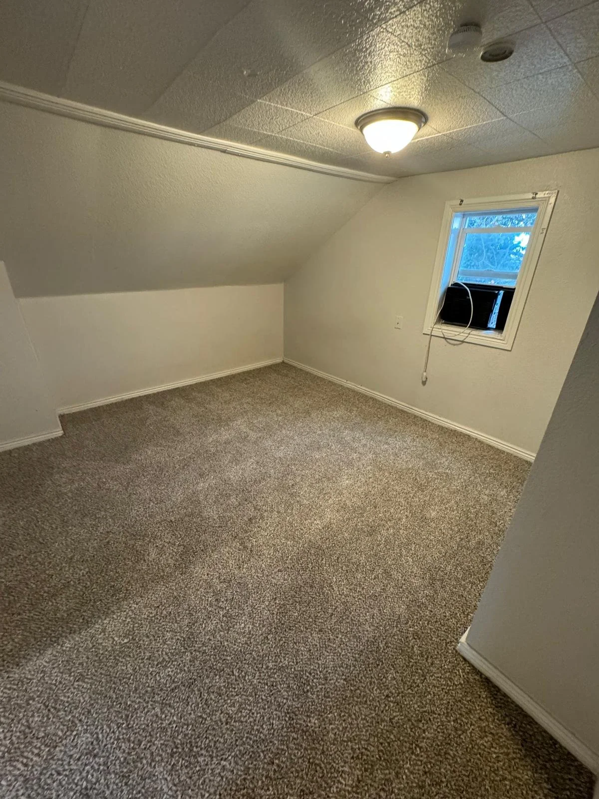 Empty room with beige carpet, a small window with black air conditioner, white walls, and ceiling light.