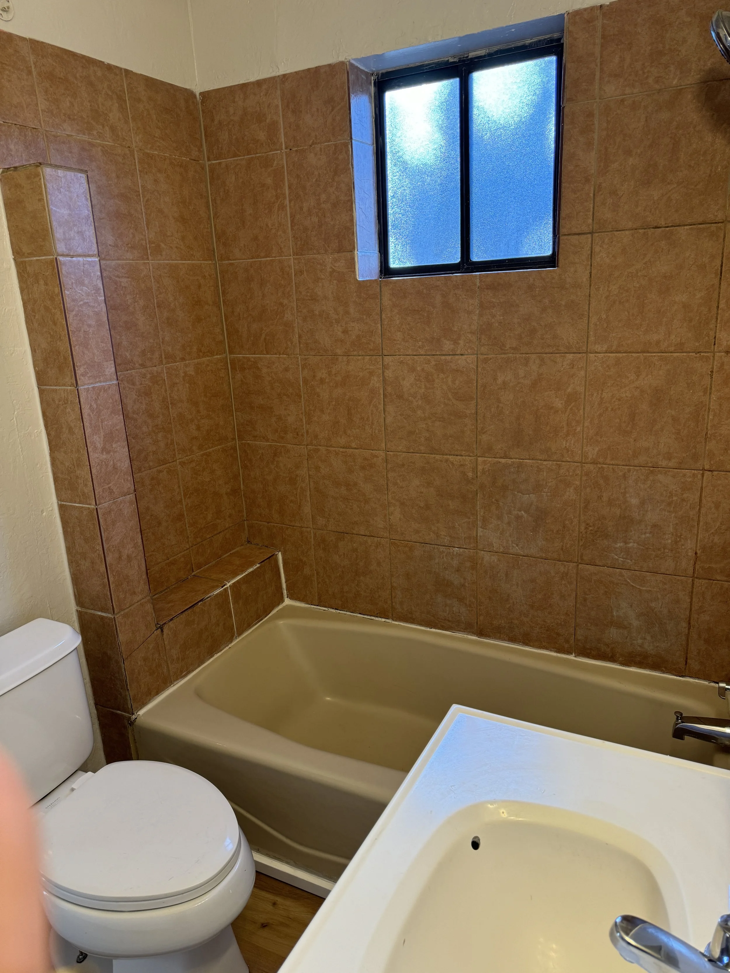 A small bathroom featuring a bathtub, a toilet, a beige sink, and a frosted window above the tub. The walls are covered with brown tiles.