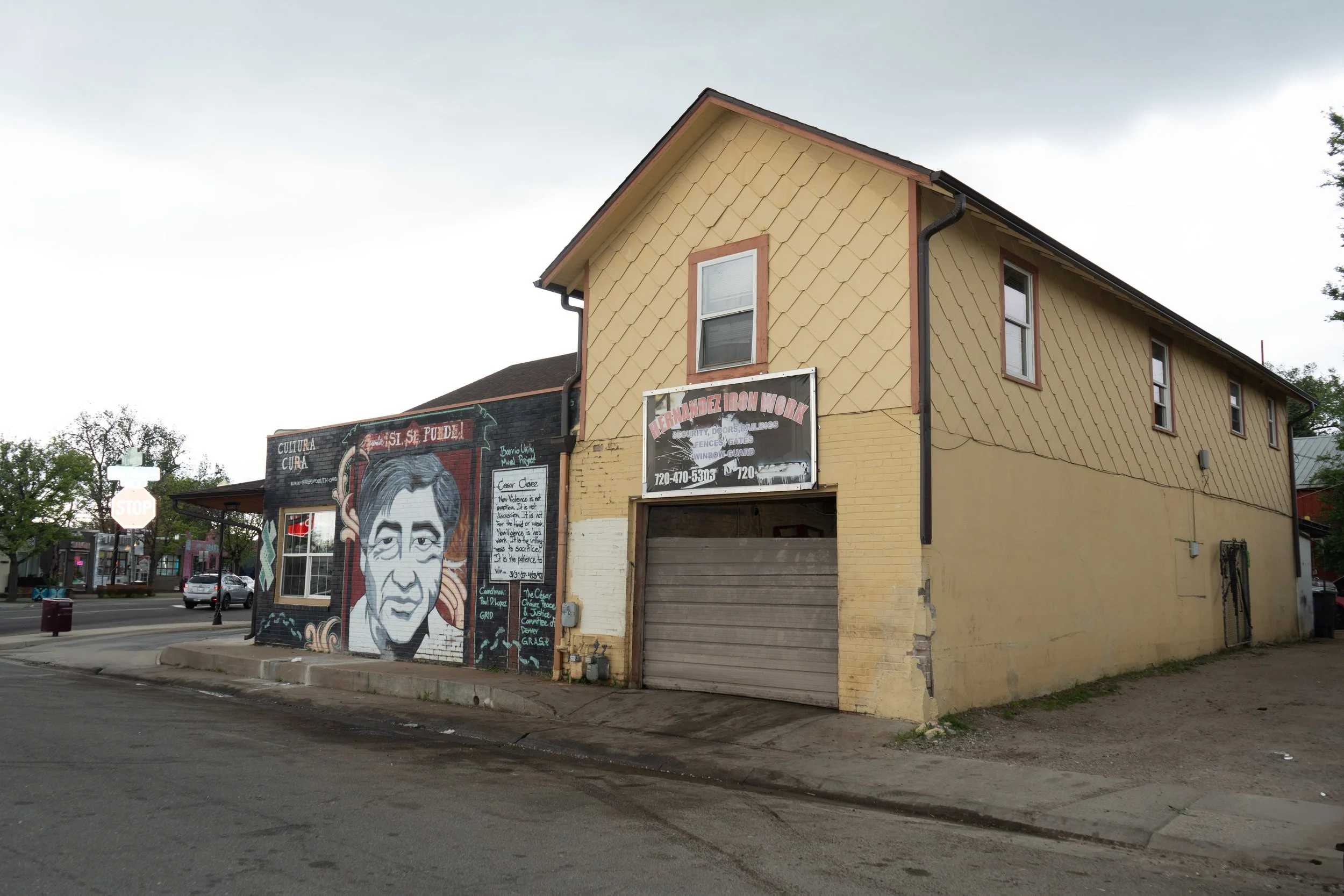 A corner building with a mural of a man's face on the side wall, next to a yellow house with a garage door, a sign for Fernandez Iron Work, and a street with cars and a stop sign.