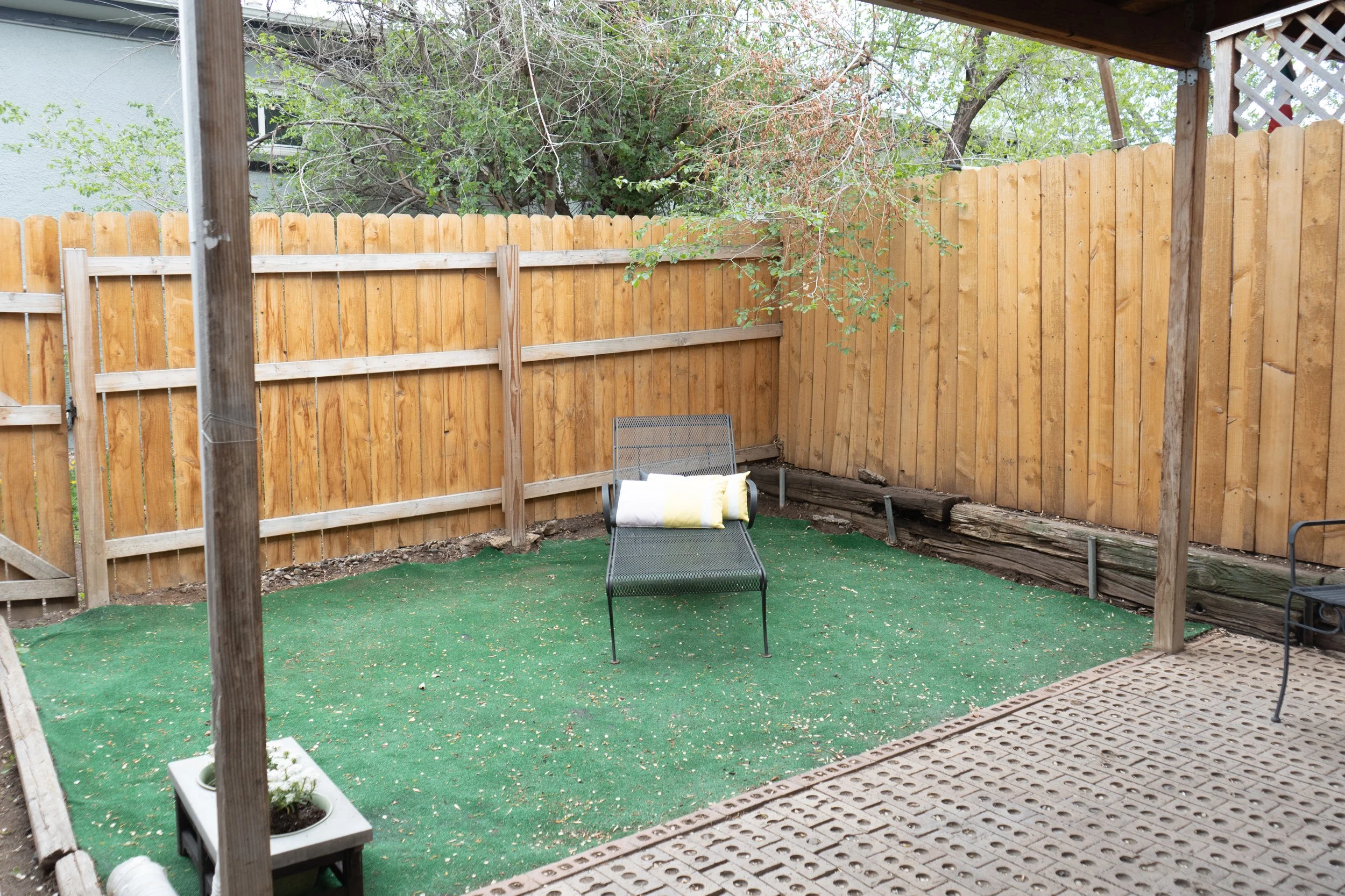 Empty backyard patio with a green artificial grass area, a black metal chair with a rolled-up towel on it, and a wooden privacy fence, partially covered by a wooden roof with trees in the background.