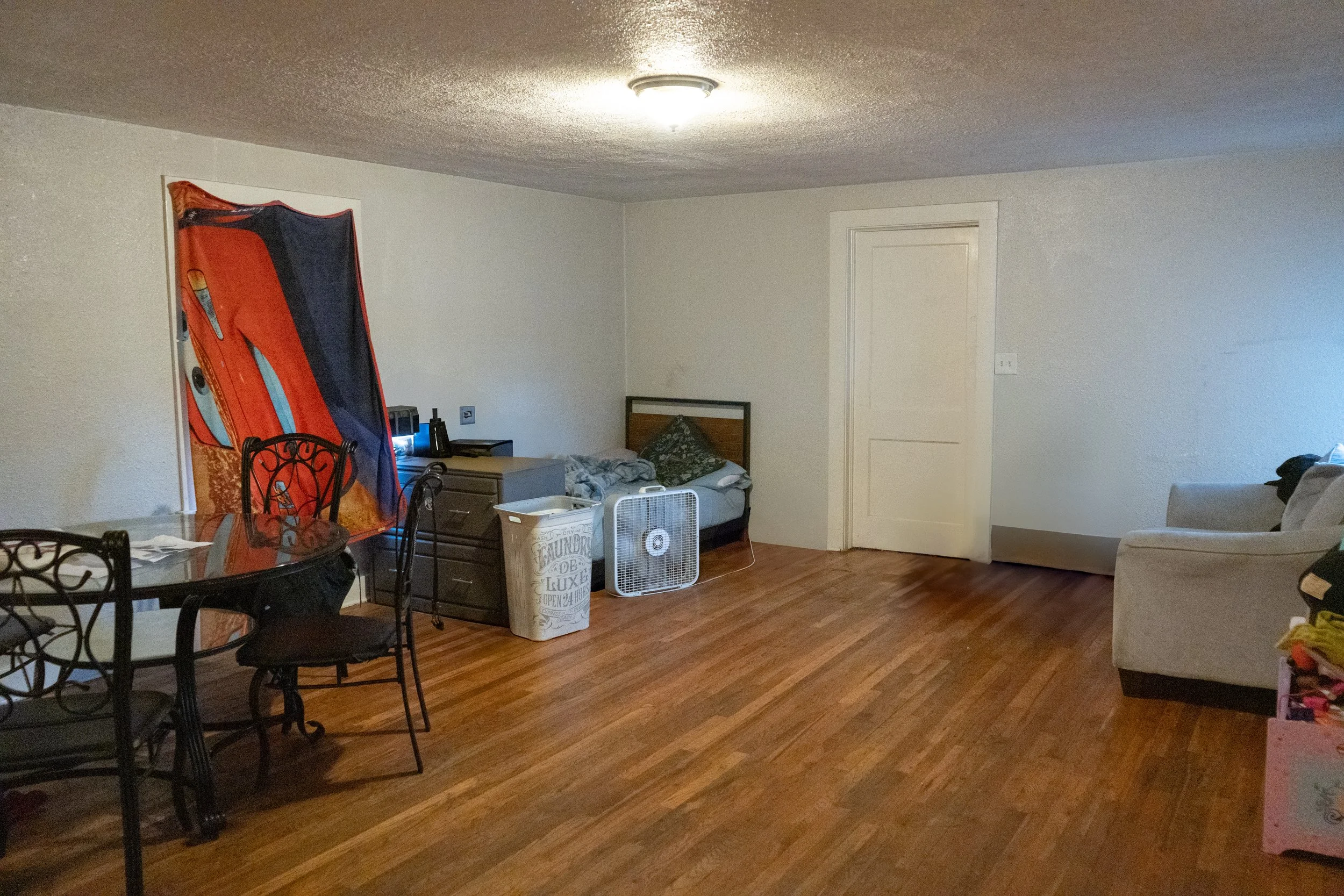Living room with hardwood floor, white walls, a dining table with chairs on the left, a bed with pillows near the center, a fan, laundry basket, and a white couch on the right. There is a window with a colorful curtain, a closed door, and a ceiling l