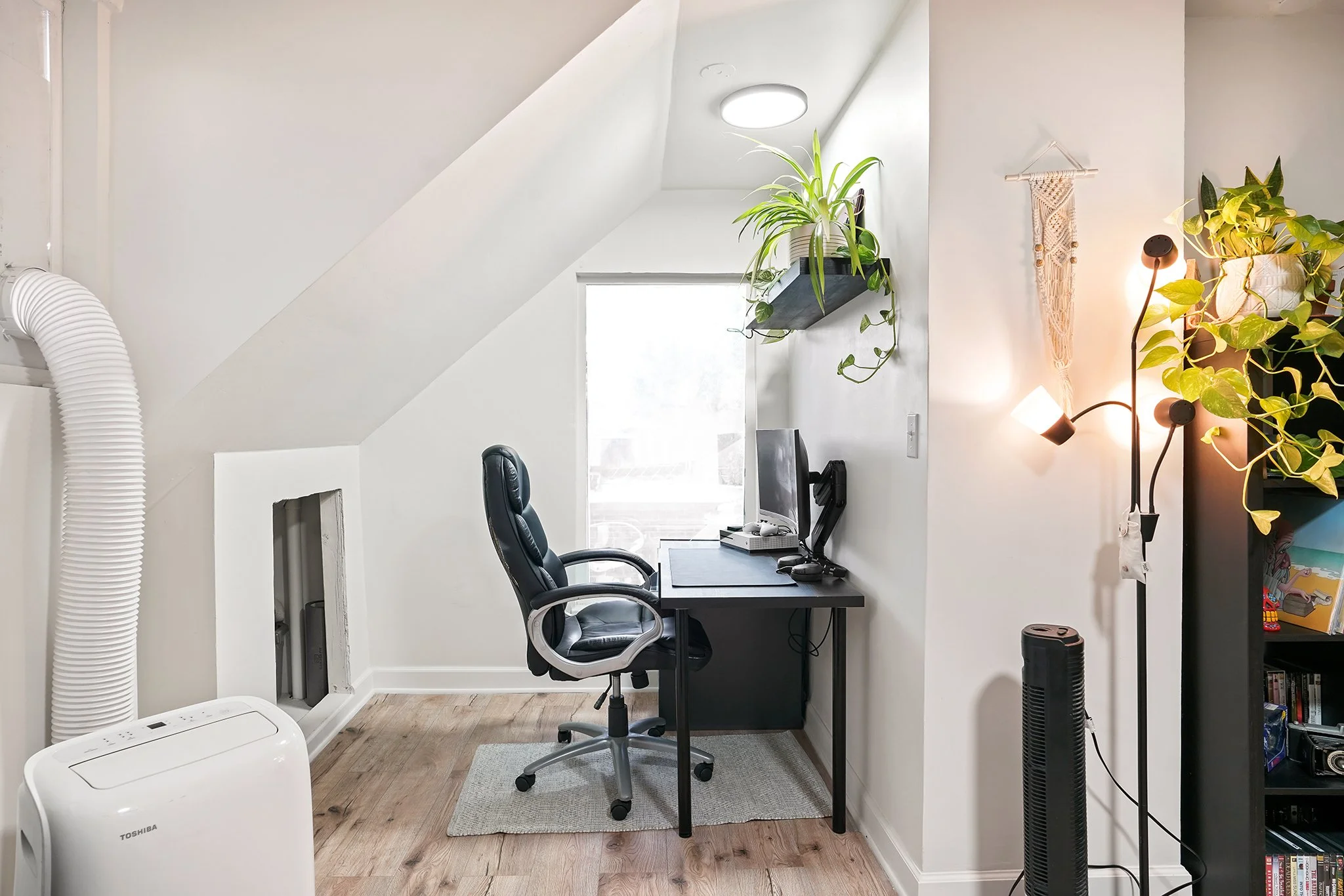 A small home office setup under a sloped ceiling, featuring a black desk with a computer monitor, a black ergonomic office chair, and two potted plants on a shelf and wall. There is a small gray rug under the chair, and a white air purifier to the le
