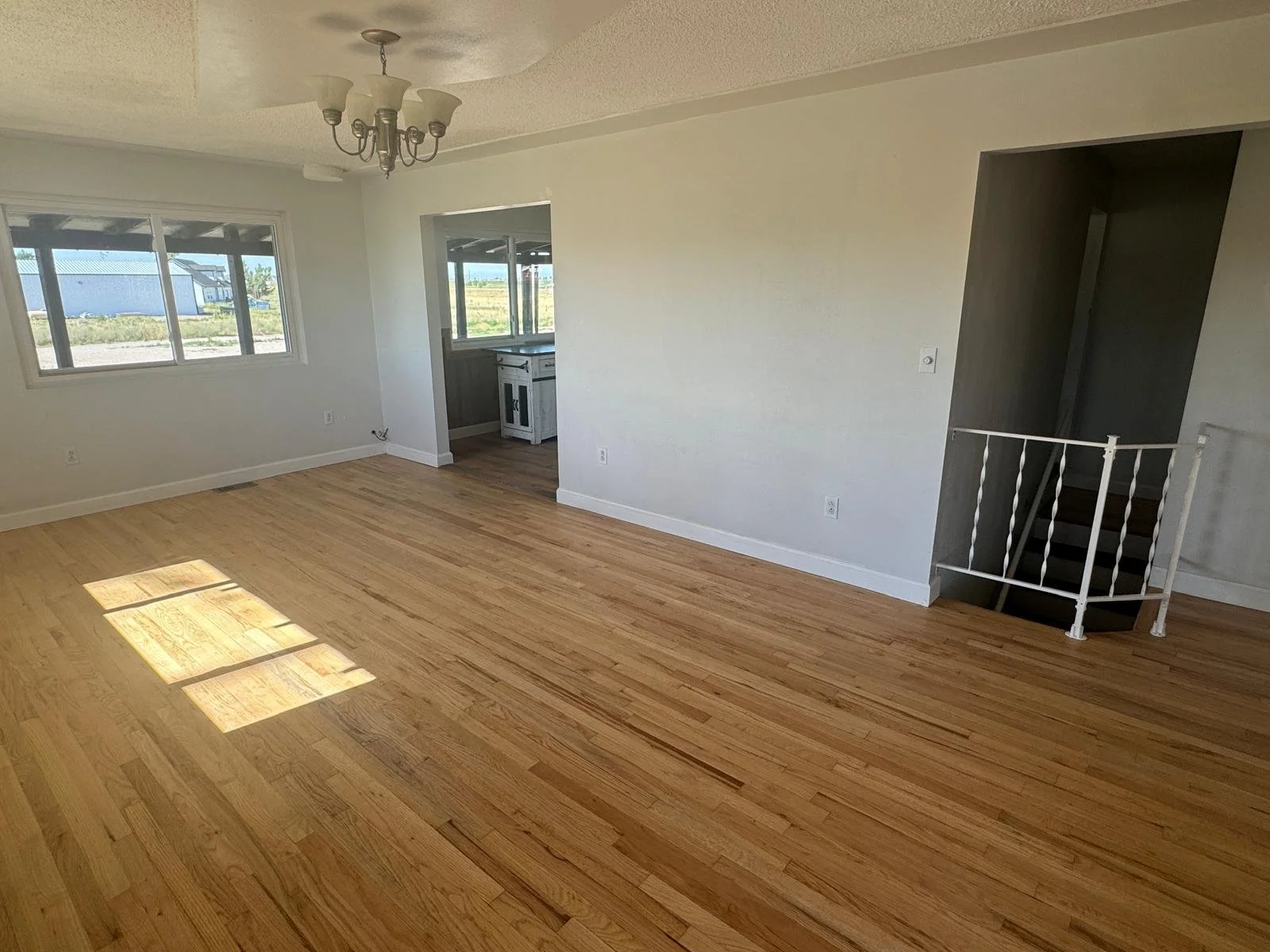 Empty living room with wood floors, white walls, large windows, a ceiling light fixture, and a staircase with a white railing.