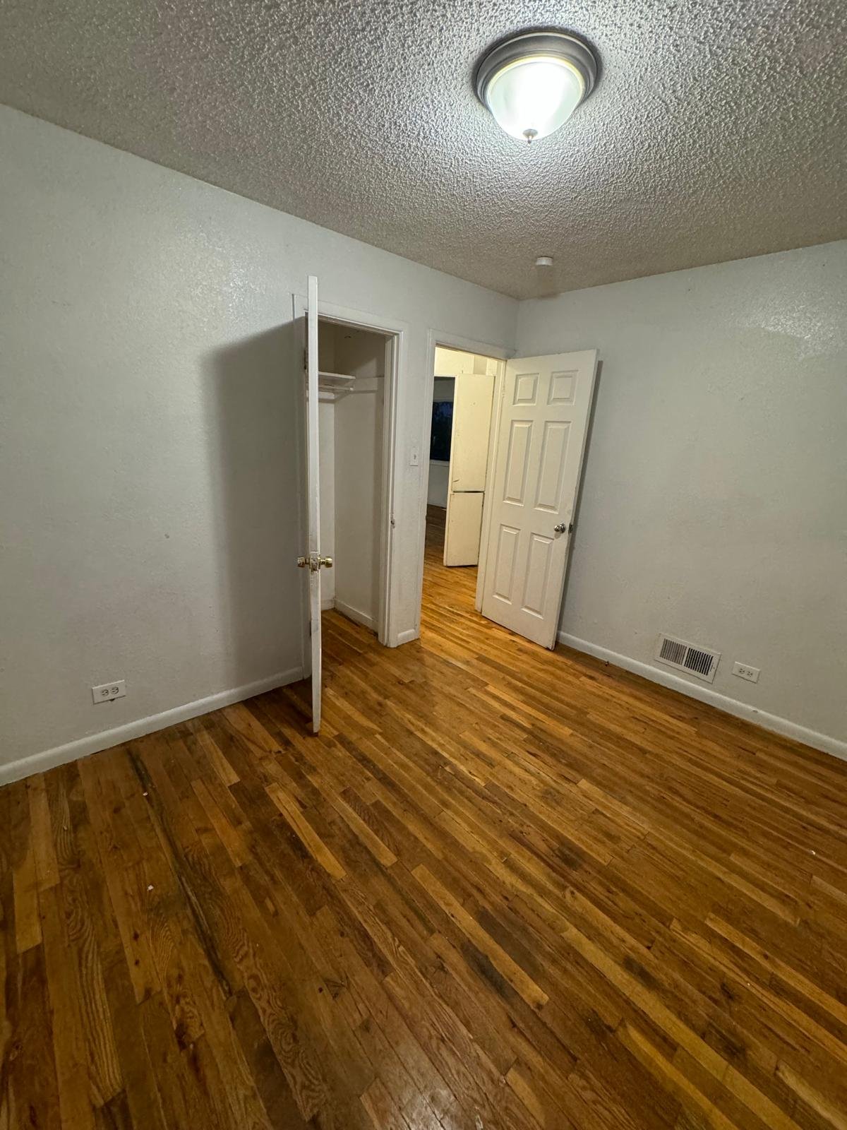 Empty room with wooden floor, white walls, a ceiling light, an open closet door, and a partially open door leading to another room.