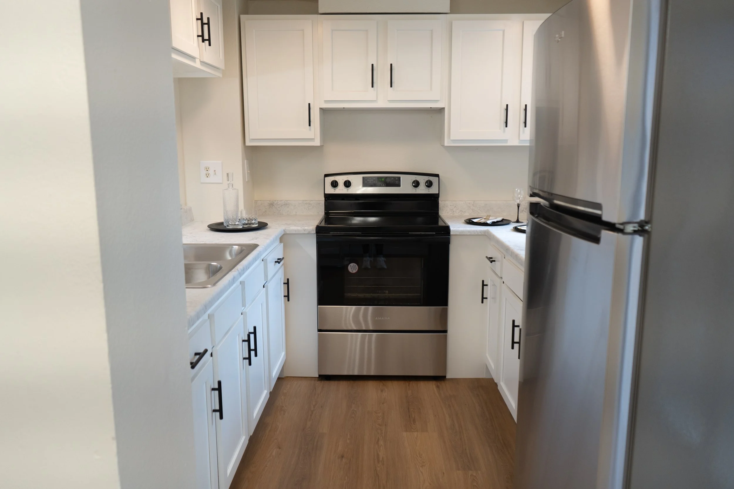 Kitchen with white cabinets, stainless steel refrigerator and oven, white countertops, and a small sink. Decor includes a tray with a glass bottle, glasses, and black plates, with a knife and fork on a napkin.