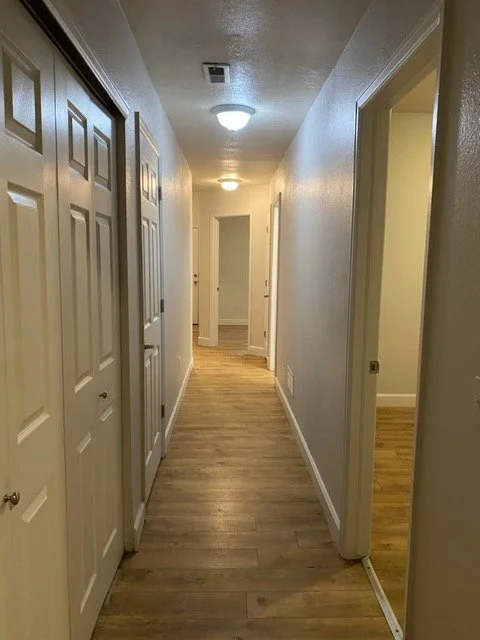 Interior of a hallway with white walls, wooden flooring, and multiple doors on each side, illuminated by ceiling lights.