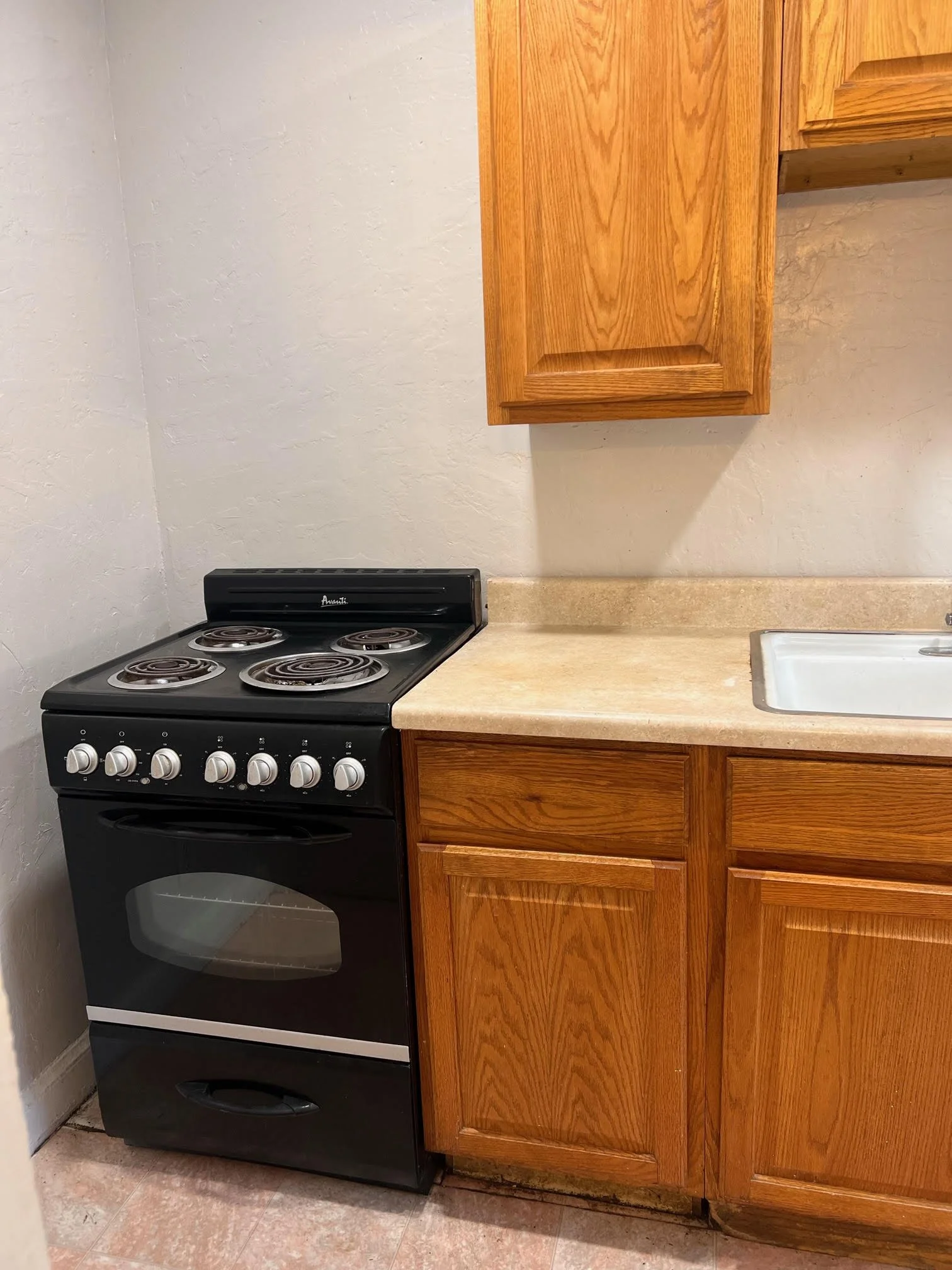 Black stove with four coil burners next to wooden kitchen cabinets and beige countertop, with white wall and sink in background.