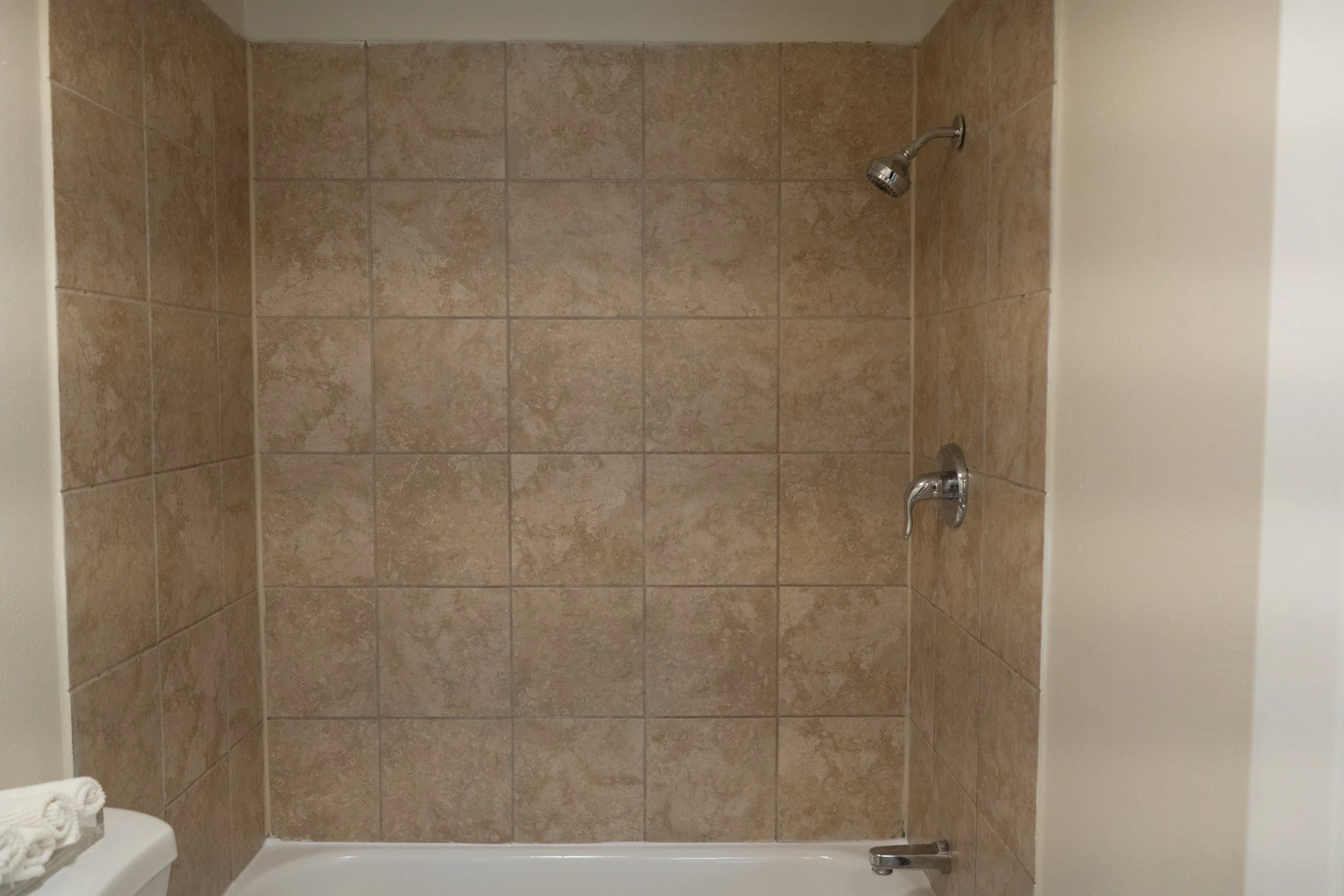 A shower with beige tiled walls, a silver showerhead, and a silver faucet, with a white bathtub base.