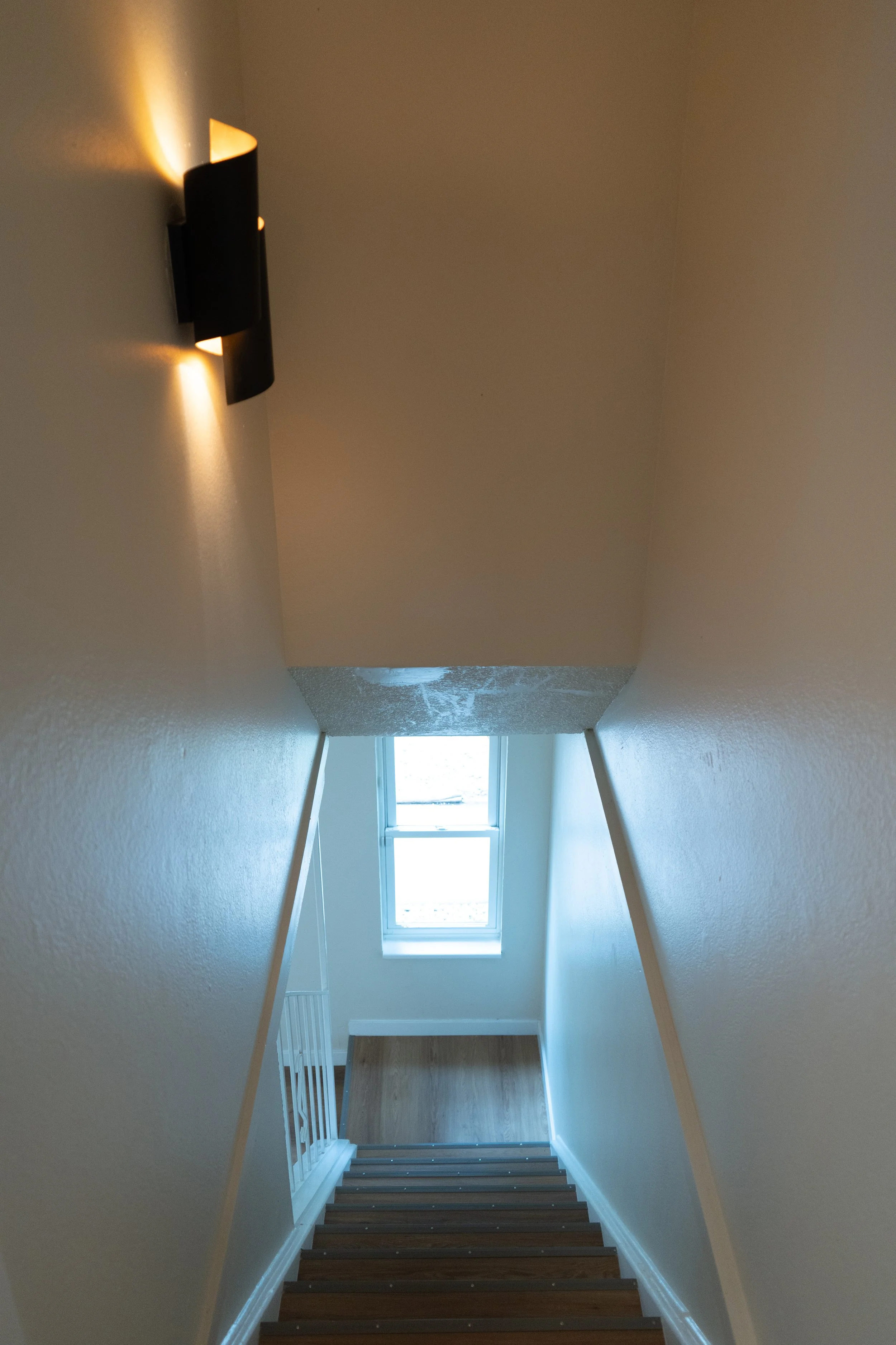 Inside view of a staircase looking down to a small landing with a window, with a wall-mounted light fixture on the left wall.