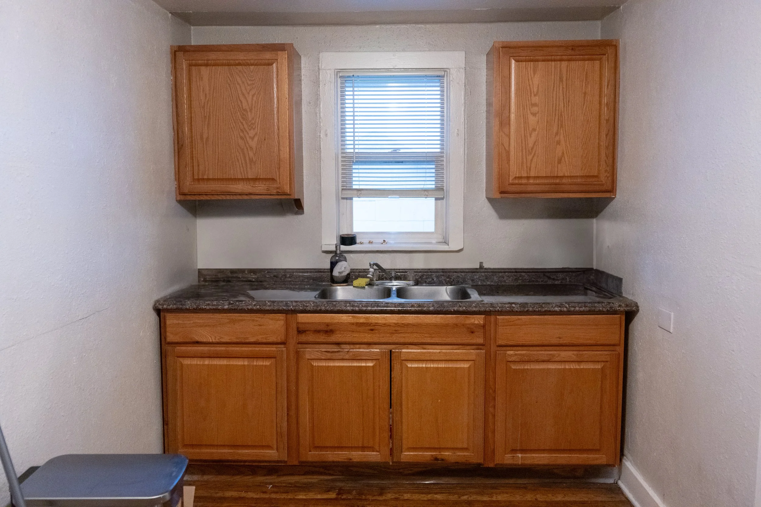 Small kitchen with wooden cabinets, granite countertop, and a window above the sink with blinds.