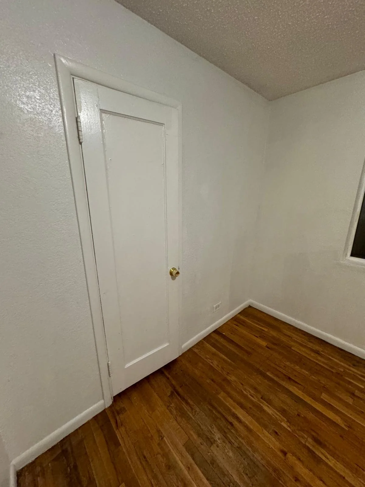 Empty room with white walls, a small white door with a gold knob, and wooden flooring.