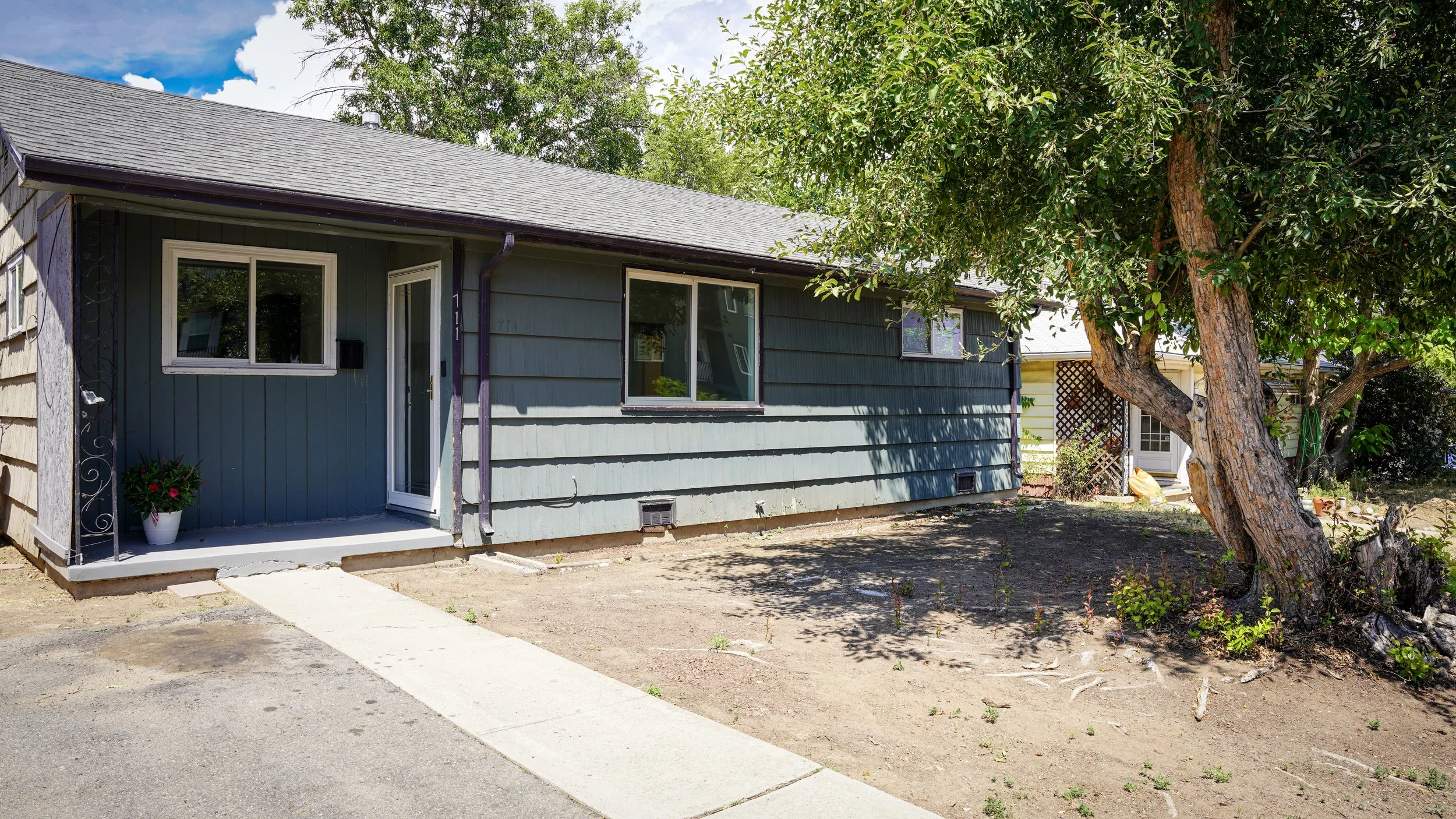 A house with blue wooden siding, a small front porch with a flower pot, and a tree with large branches in the front yard. The yard is mostly bare soil, and there is a sidewalk leading to the front door.