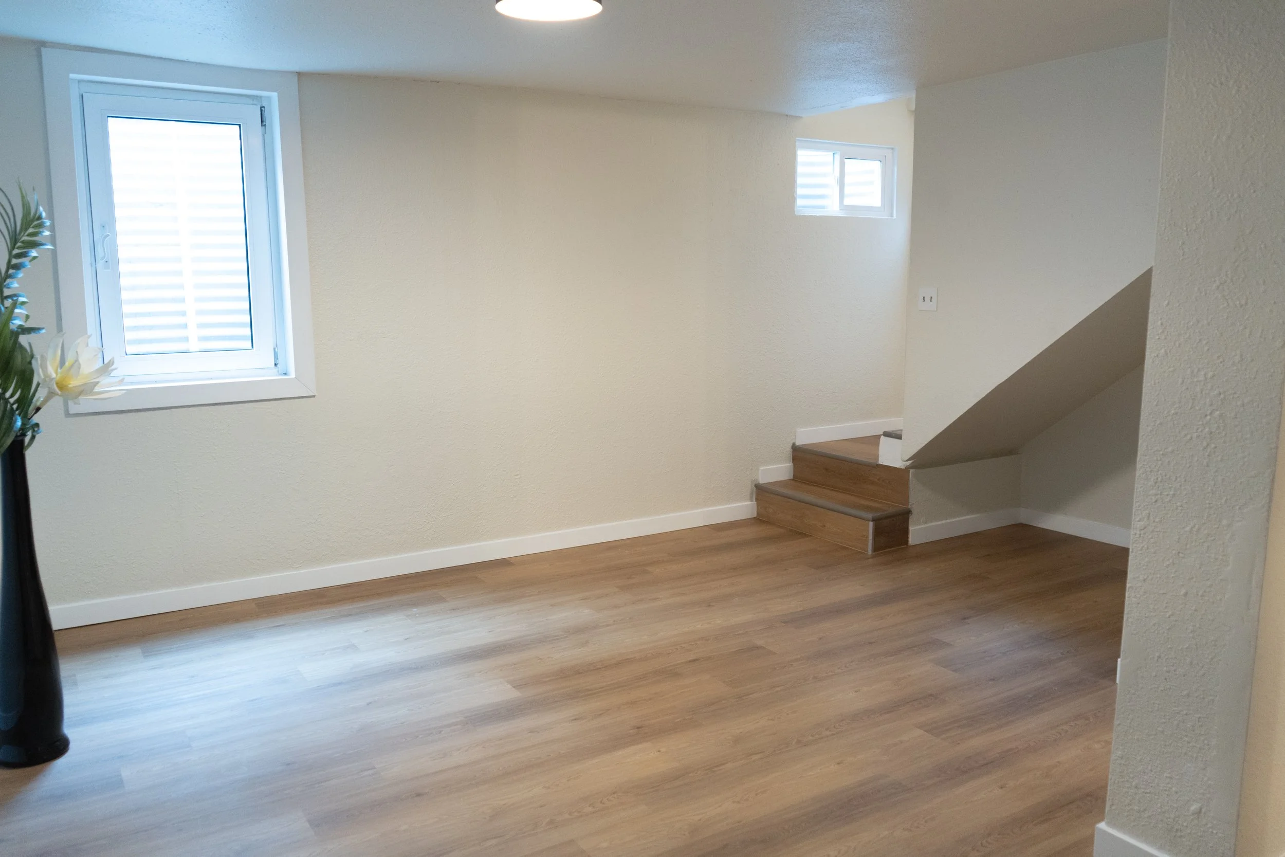 Empty room with light-colored walls, wooden flooring, two small windows, and a staircase with wooden steps.