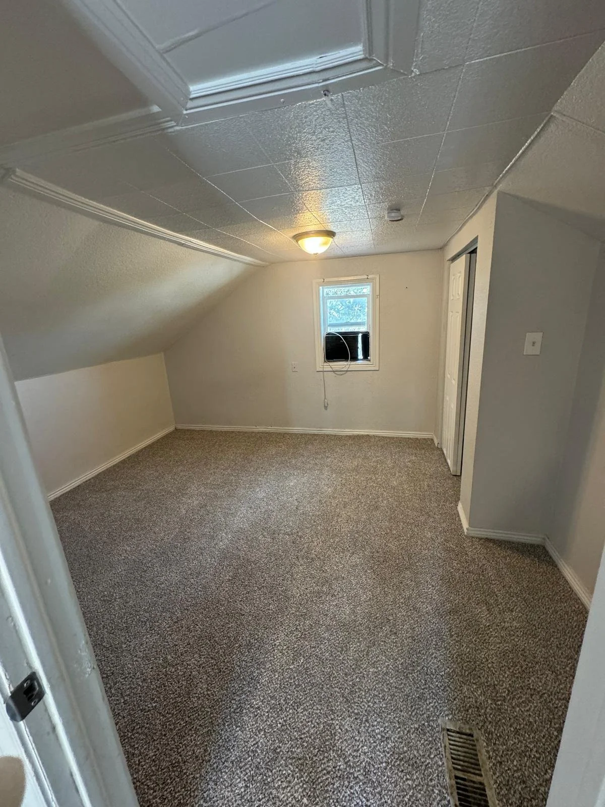 Empty small room with sloped ceiling, beige carpet, white walls, a small window with black objects on the windowsill, and a closed white closet door.