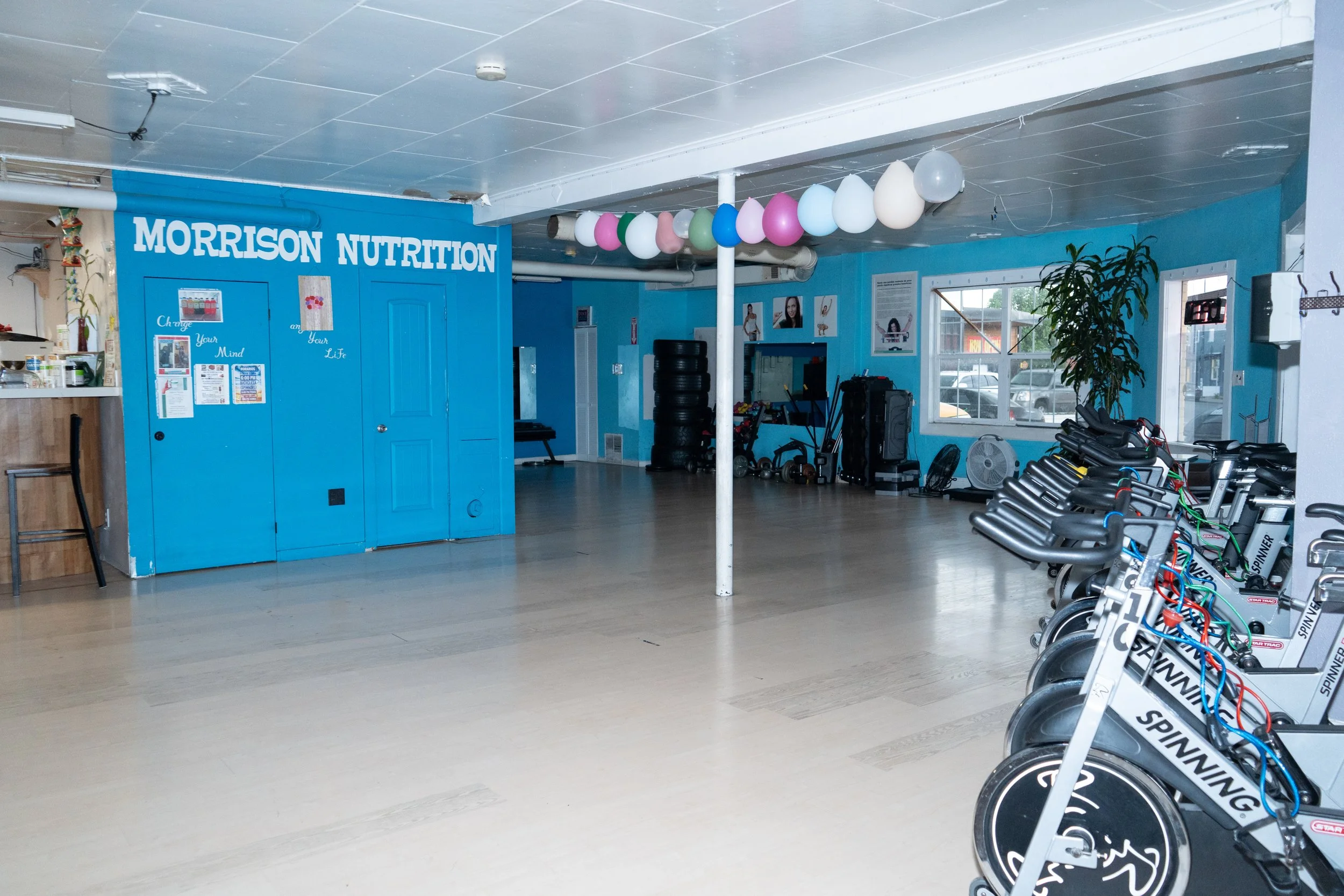 Empty fitness studio with stationary bikes lined up on the right, balloons hanging from the ceiling, and blue walls with the sign 'Morrison Nutrition' on the left.