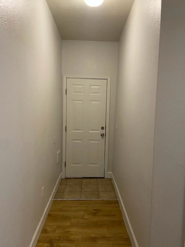 Narrow entryway with white walls, ceiling light fixture, white door at the end, tiled floor area just inside the door, and wood flooring leading up to it.