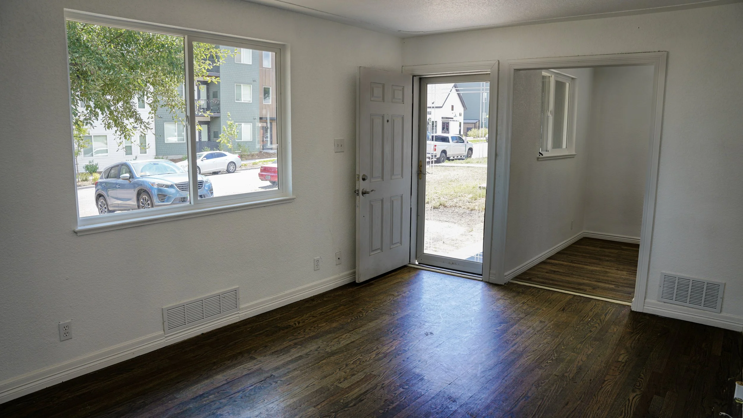 Empty room with large window and glass door leading outside. Wooden floor, white walls, small room with window visible through an open doorway.
