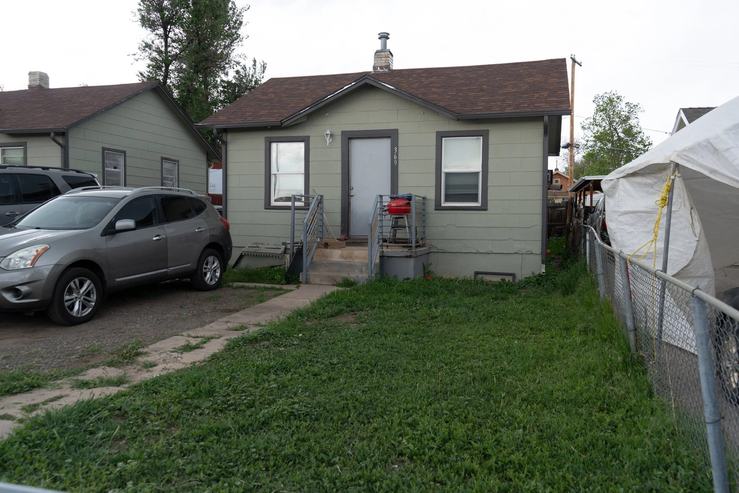 A small, light-green house with a brown roof, front steps, and a white door. Two windows are on either side of the door. A car is parked to the left, and a white tent or canopy is on the right, with a chain-link fence running alongside it. Green gras