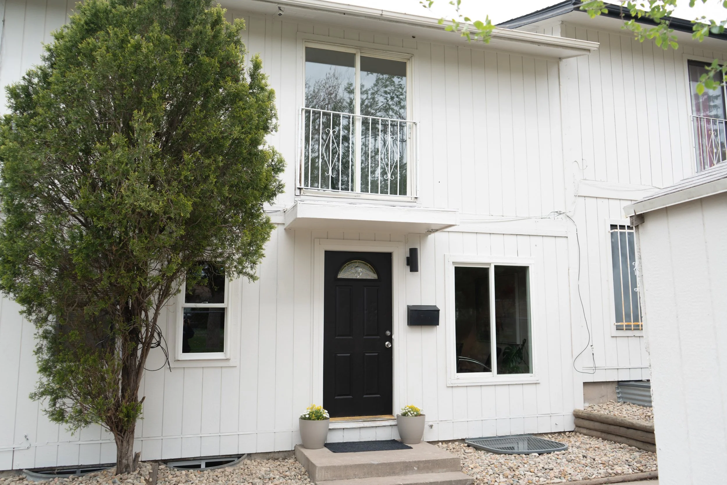 Front view of a white two-story house with a black front door, a small balcony above, two potted plants near the door, and a tree on the left side.
