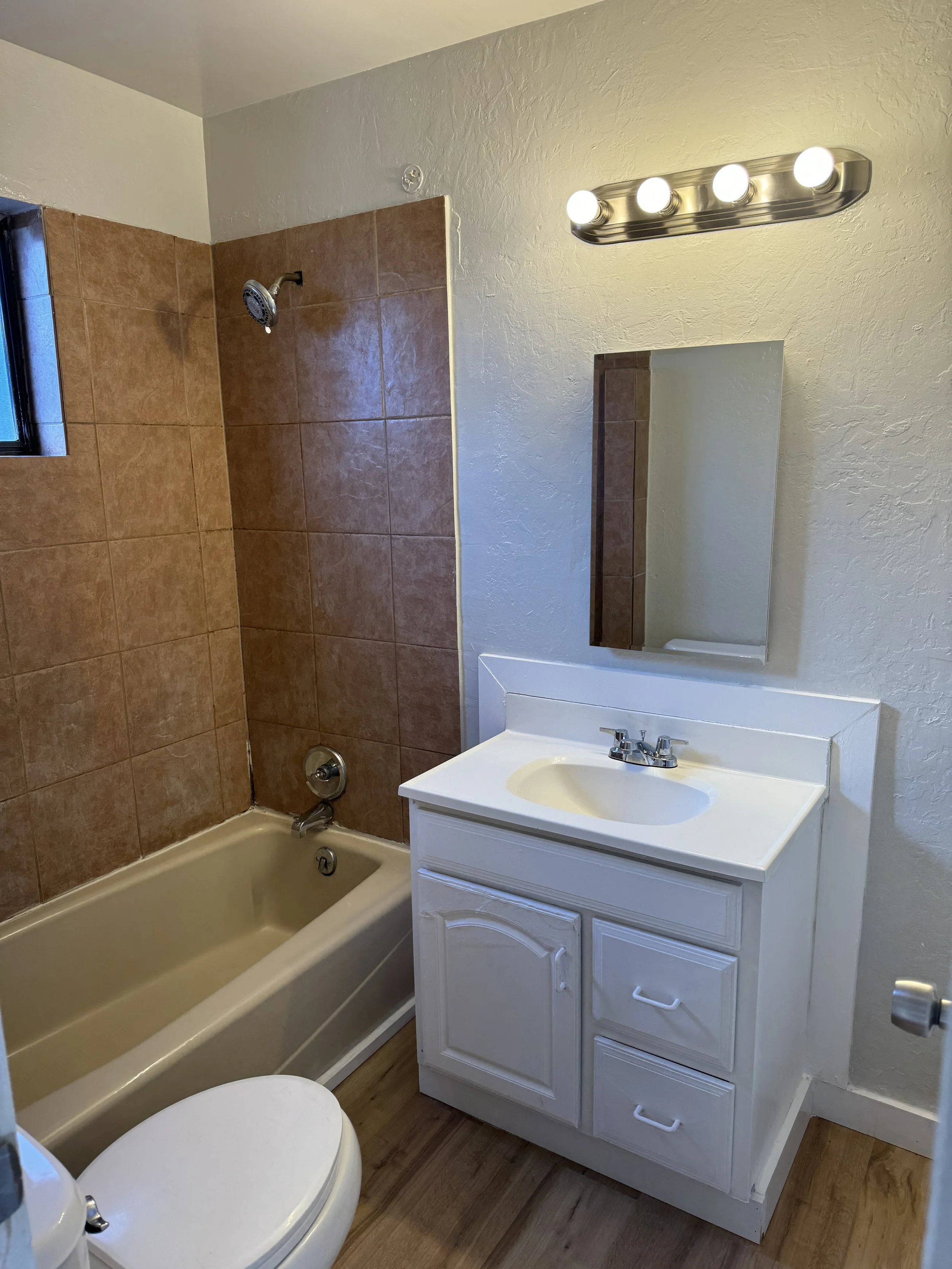A small bathroom featuring a bathtub with brown tiled walls, a small window, a white vanity with a mirror, and a four-bulb light fixture above the mirror.