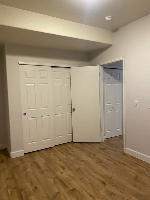 Empty room with hardwood floors, white walls, a small closet with sliding doors, and an open door leading to another room.