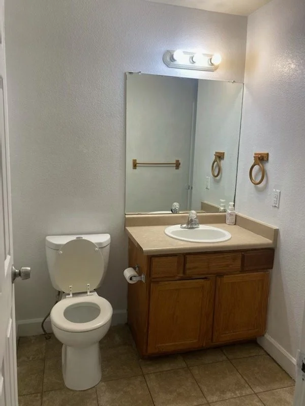 Bathroom with toilet and wooden vanity with beige countertop, mirror, and wall-mounted towel rings.