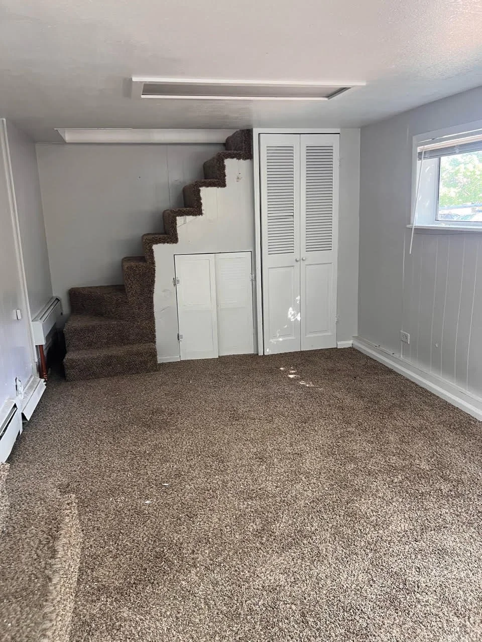 Empty room with beige carpet, white walls, window on the right, and a staircase with brown carpet leading up on the left. Small closet doors beneath the stairs and louvered closet doors to the right of the stairs.