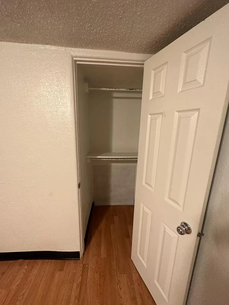 Empty closet with white door slightly open, showing two wire shelves inside, located in a room with beige walls and wooden flooring.