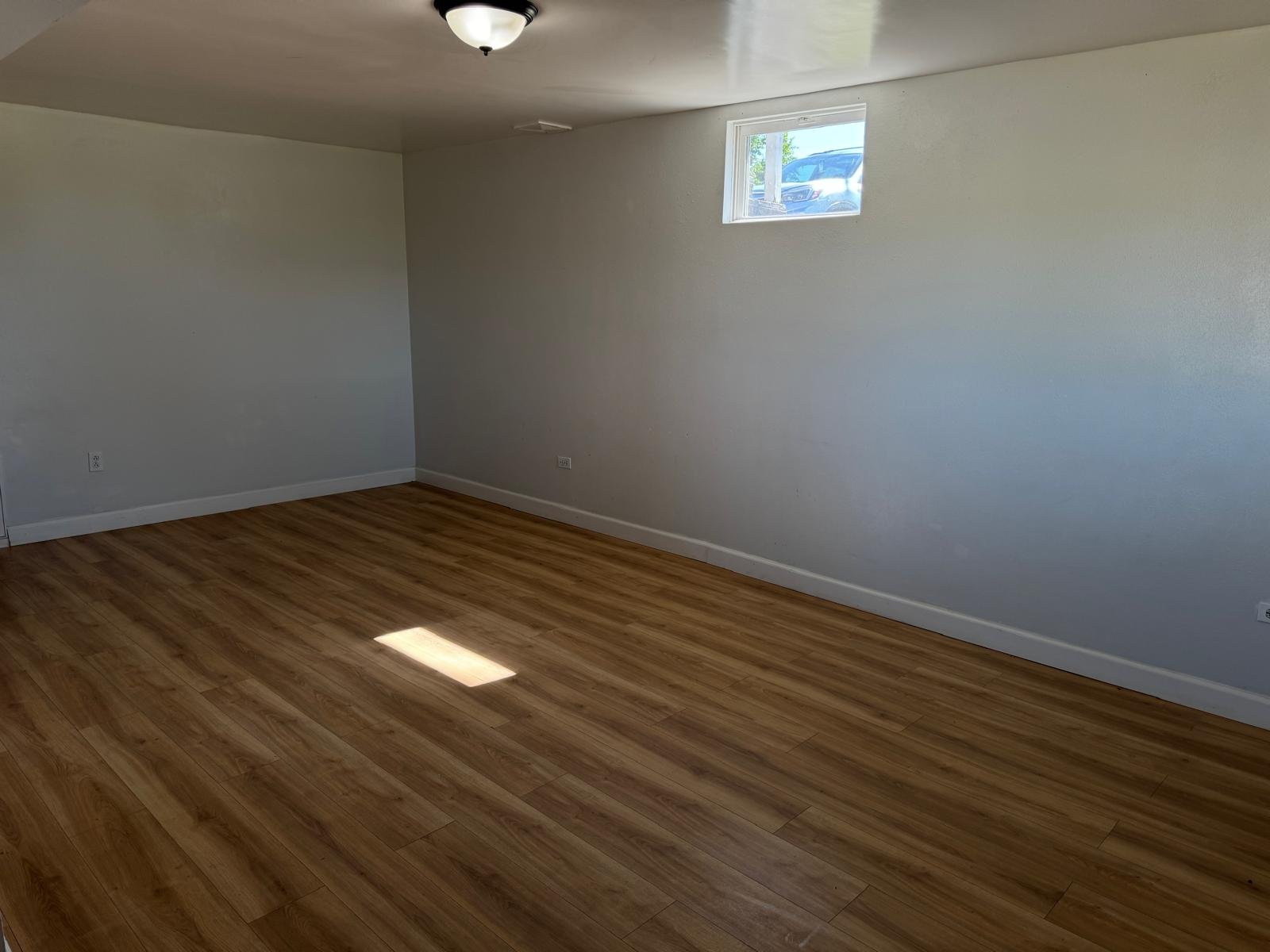 Empty room with white walls, wooden flooring, a small window, and a ceiling light fixture.