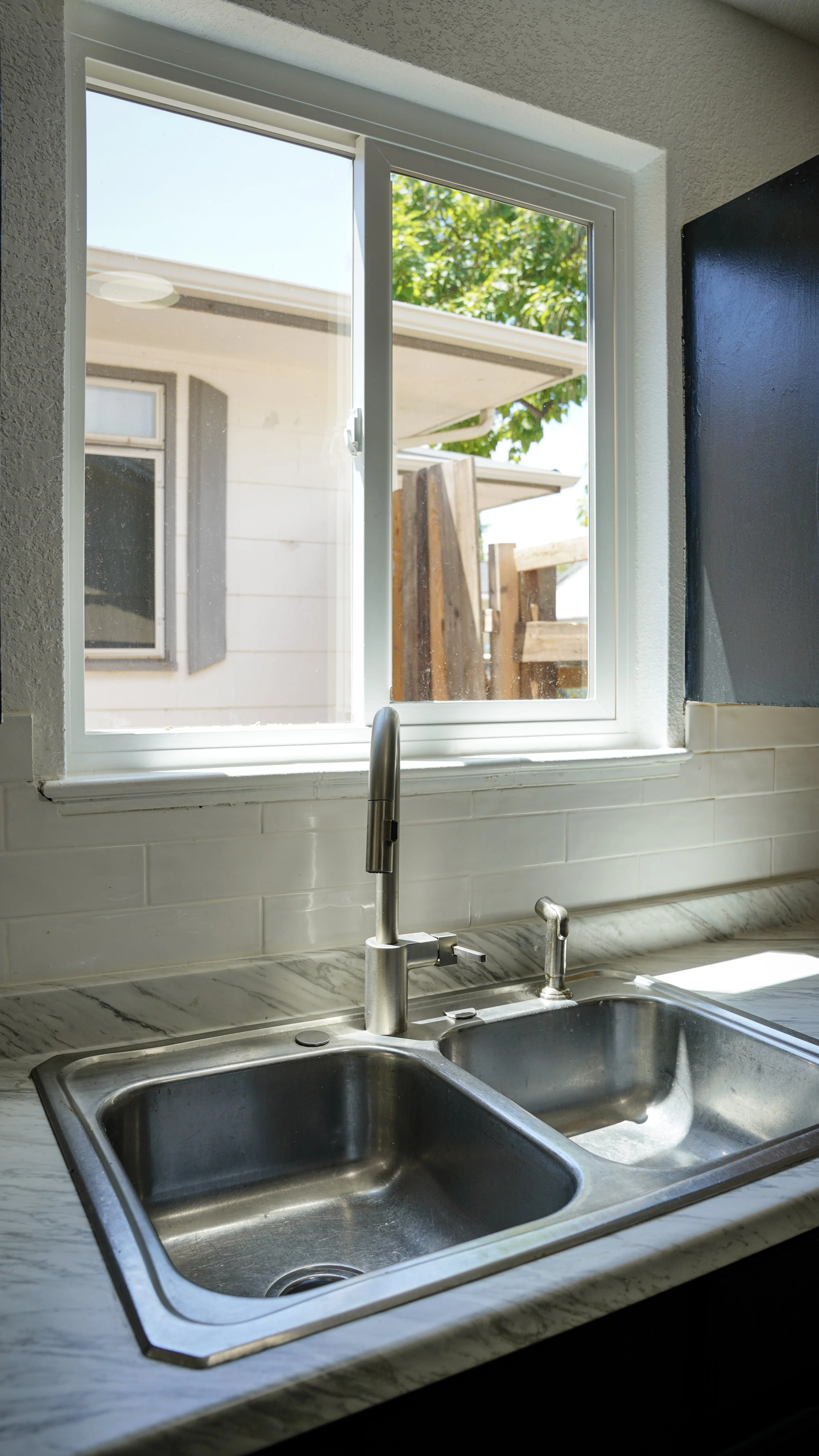 Kitchen sink with a view of a backyard through a window, showing a house, green trees, and wooden fence panels.