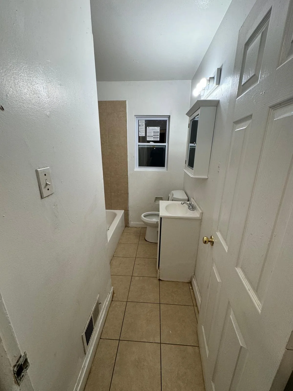 Small bathroom with beige tiled floor, white walls, a window, a bathtub, a toilet, and a small sink with a mirror cabinet.