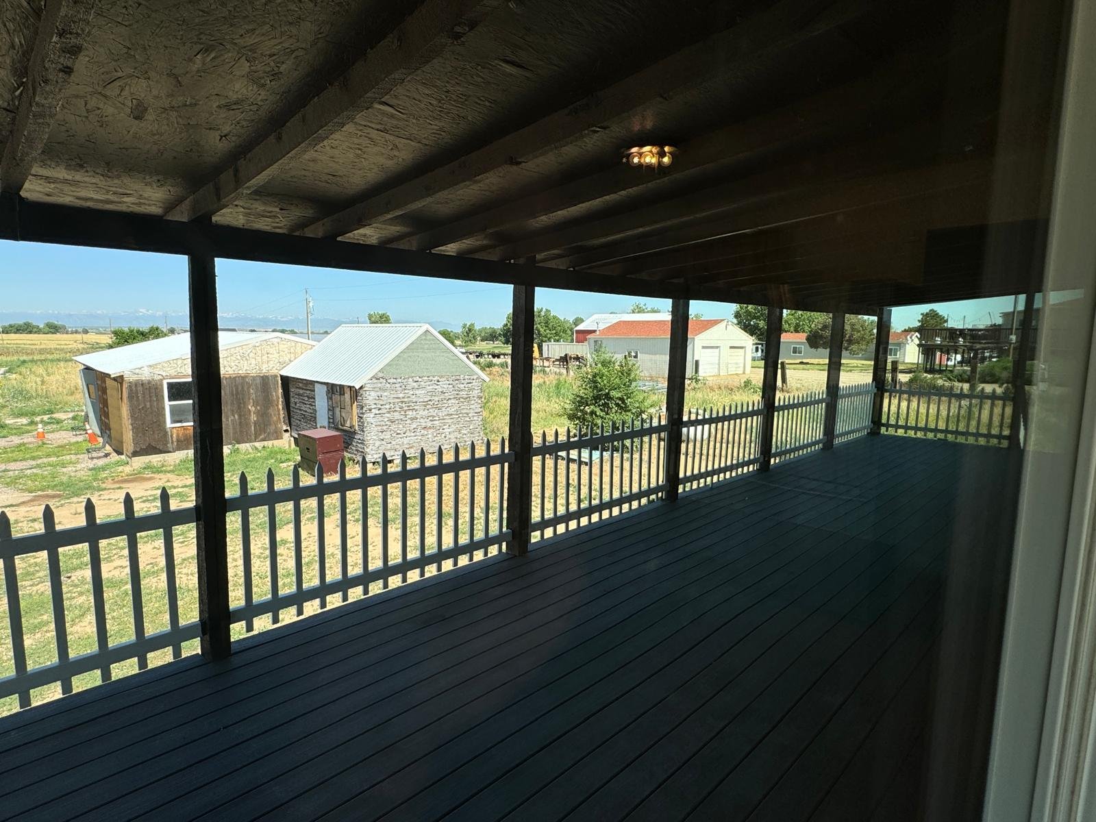 A covered porch with a wooden deck and white picket fence railing overlooking a rural area with small barns, grass, and a few houses in the distance.