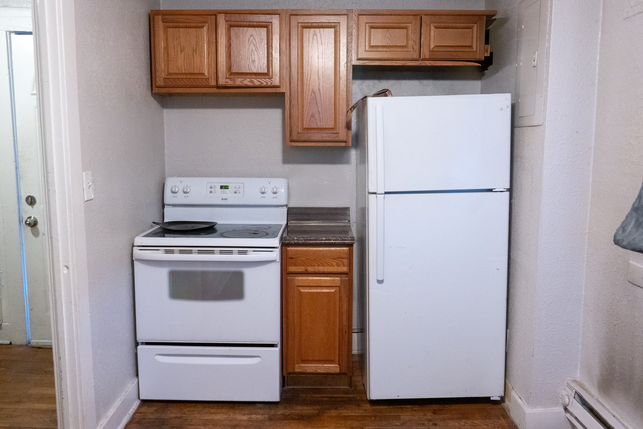 A small kitchen with wooden cabinets, a white stove with a frying pan on top, a brown countertop, and a white refrigerator. The floor is wood, and the wall is light-colored.
