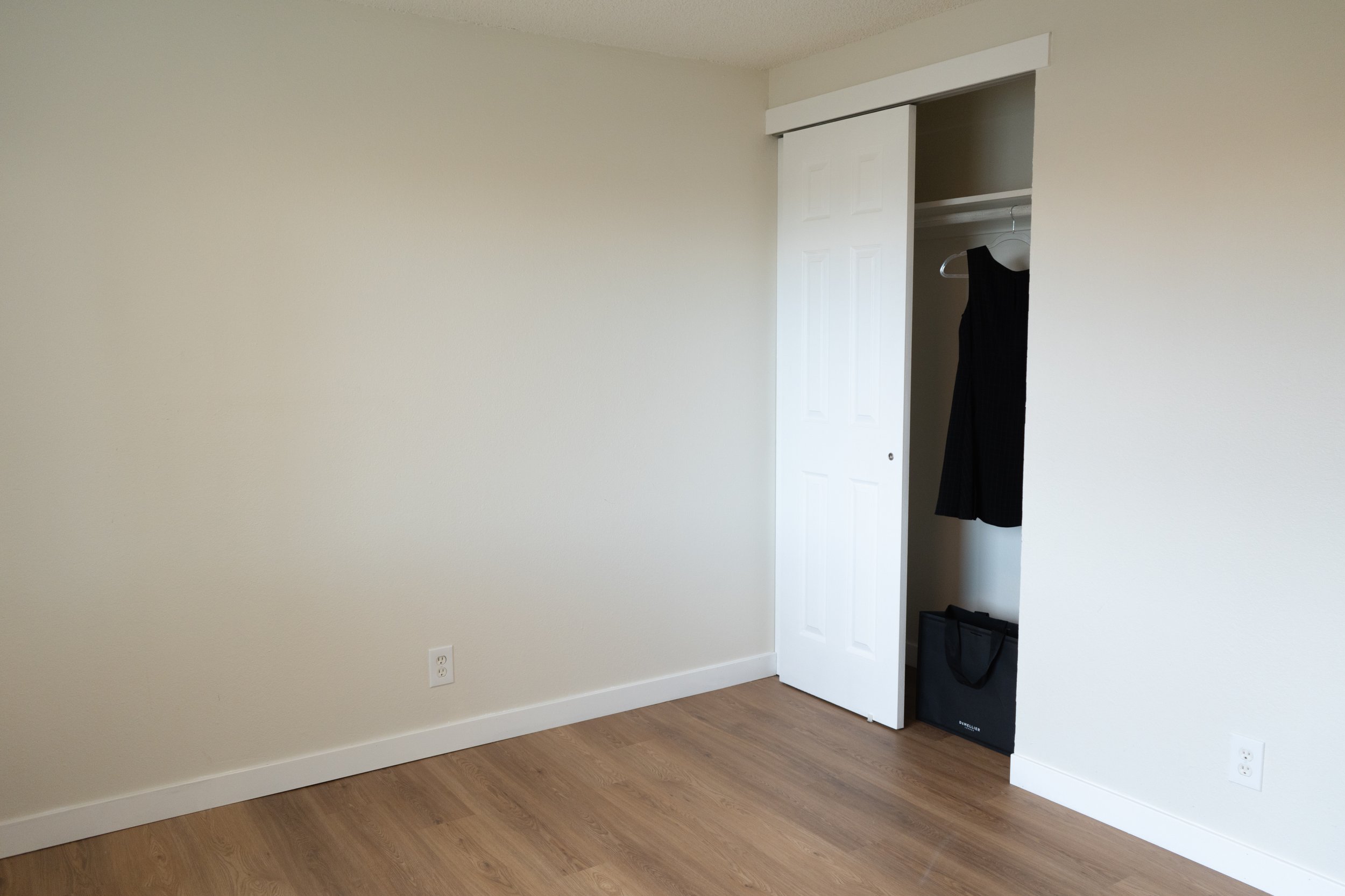 Empty room with light-colored walls, a closet with a sliding door, a black dress hanging inside, and a black bag on the floor. Wooden flooring and electrical outlets are visible.