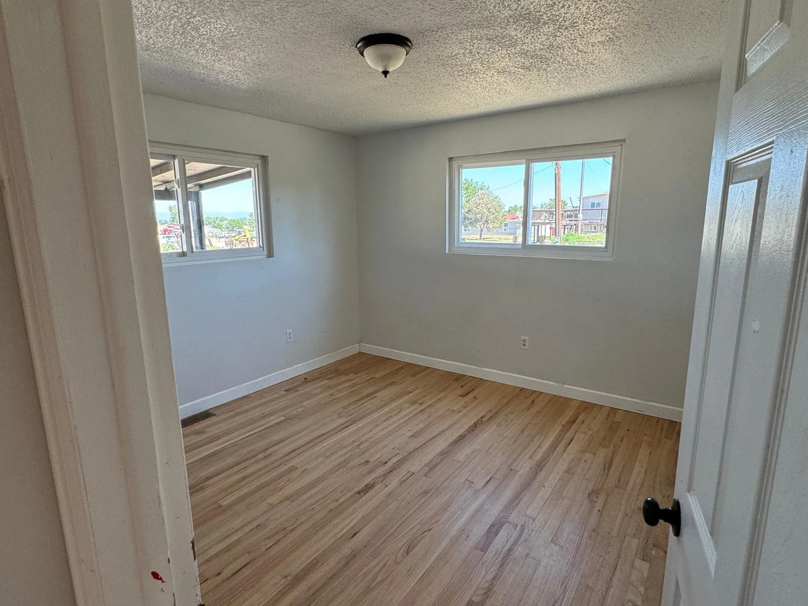 Empty room with hardwood floors, white walls, three windows, a ceiling light fixture, and a part of a door visible.