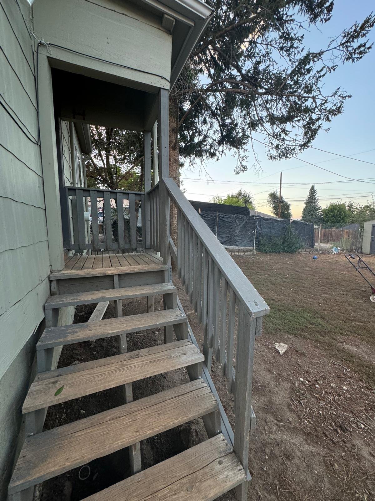 Backyard with aged wooden stairs and porch, green house wall, large trees, and a fenced yard with gardening tools and a lawnmower.