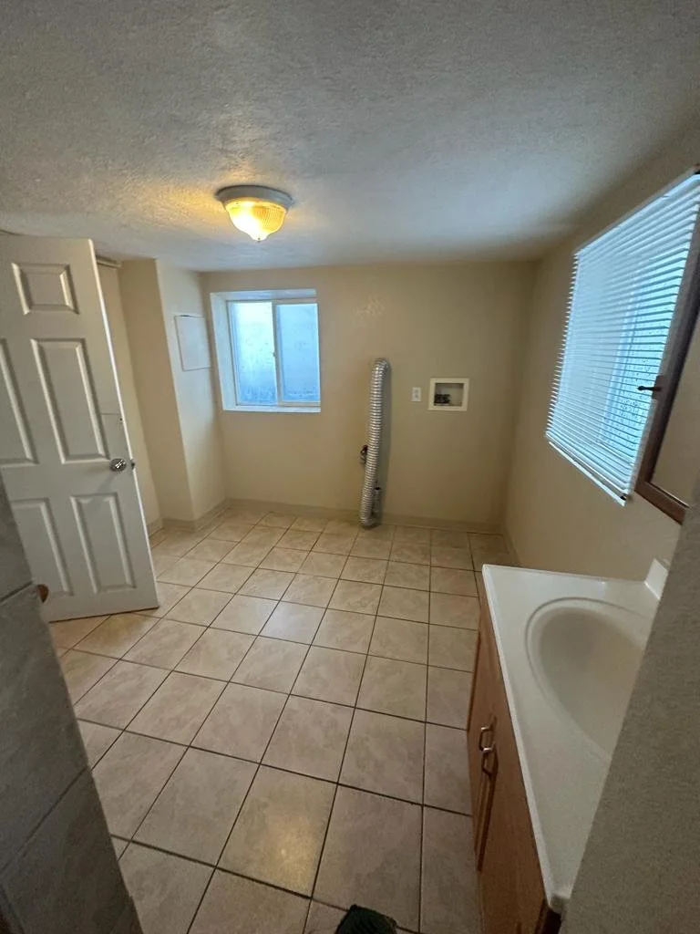 Empty laundry or utility room with tiled floor, a window, a small sink with a wooden cabinet, a ceiling light, and a vent pipe along the wall.