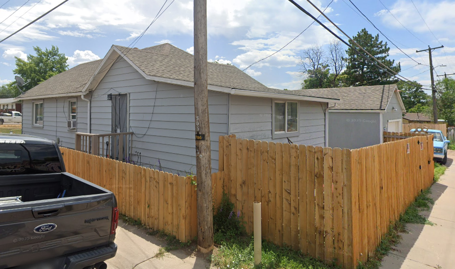 A small, single-story house with white siding, a fenced yard, and a porch with wooden steps. There is a wooden fence surrounding the yard and a utility pole in front. Several cars are parked nearby under a partly cloudy sky.