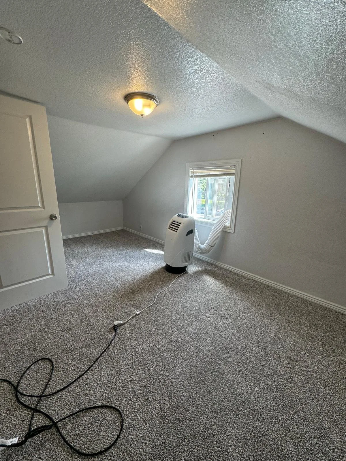 Empty room with carpeted floor, ceiling light fixture, white walls, small window, and a portable air conditioner with a hose attached.