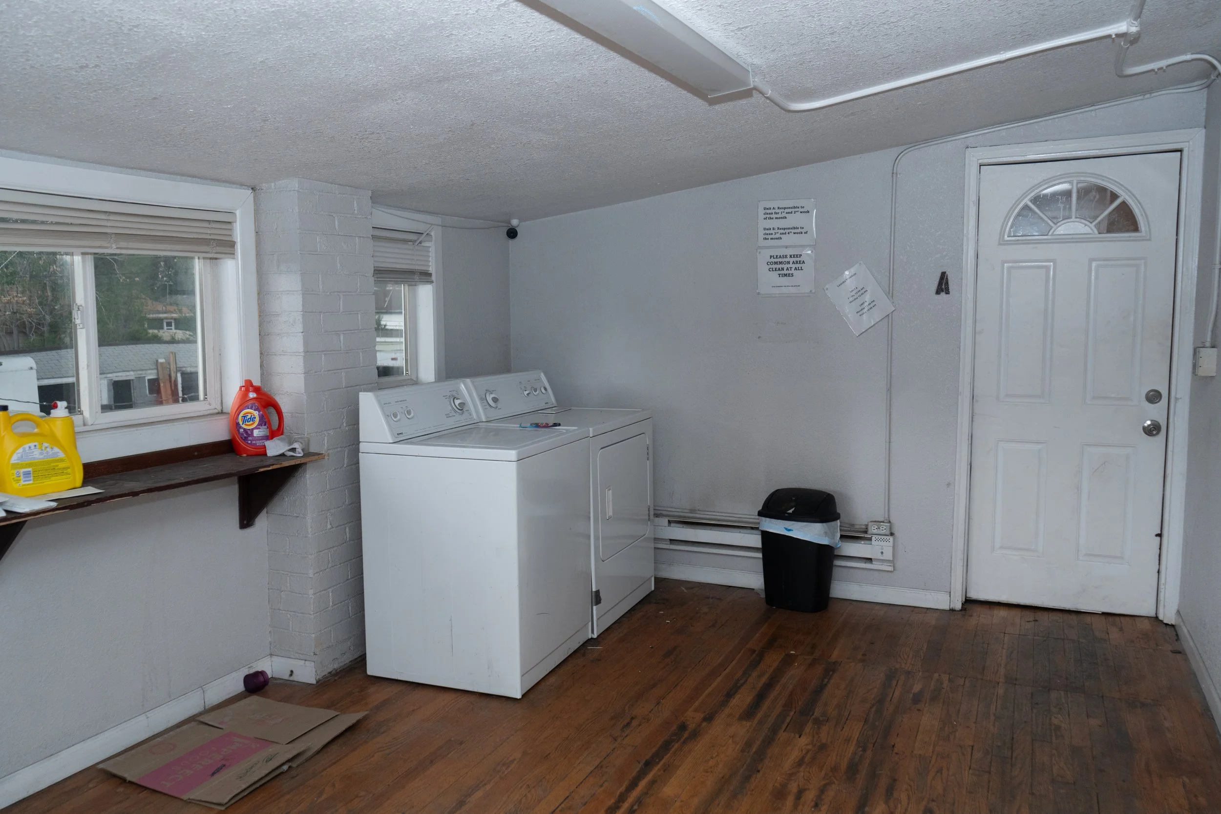 An empty laundry room with a white washing machine beside a matching dryer, a black trash bin, two windows with blinds, laundry detergent bottles on a wooden shelf, and signs on the wall near a white door.