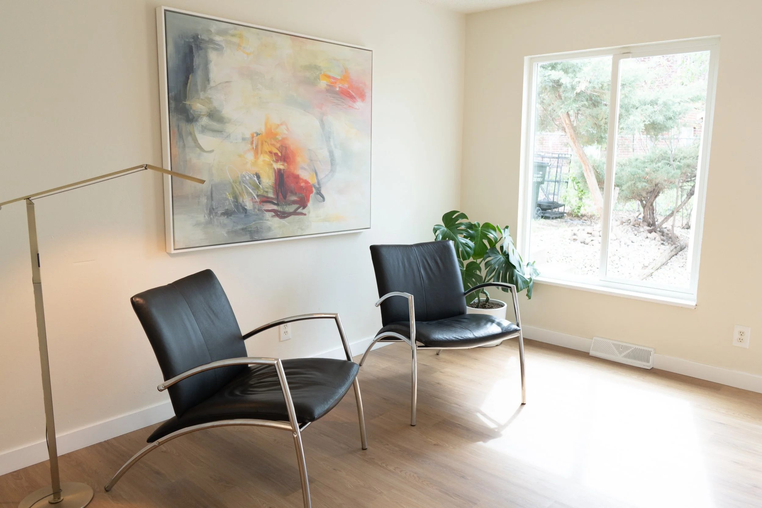Waiting room with two black leather chairs, a large abstract painting on the wall, a tall floor lamp, a potted plant near a window, and a view of a small garden outside.