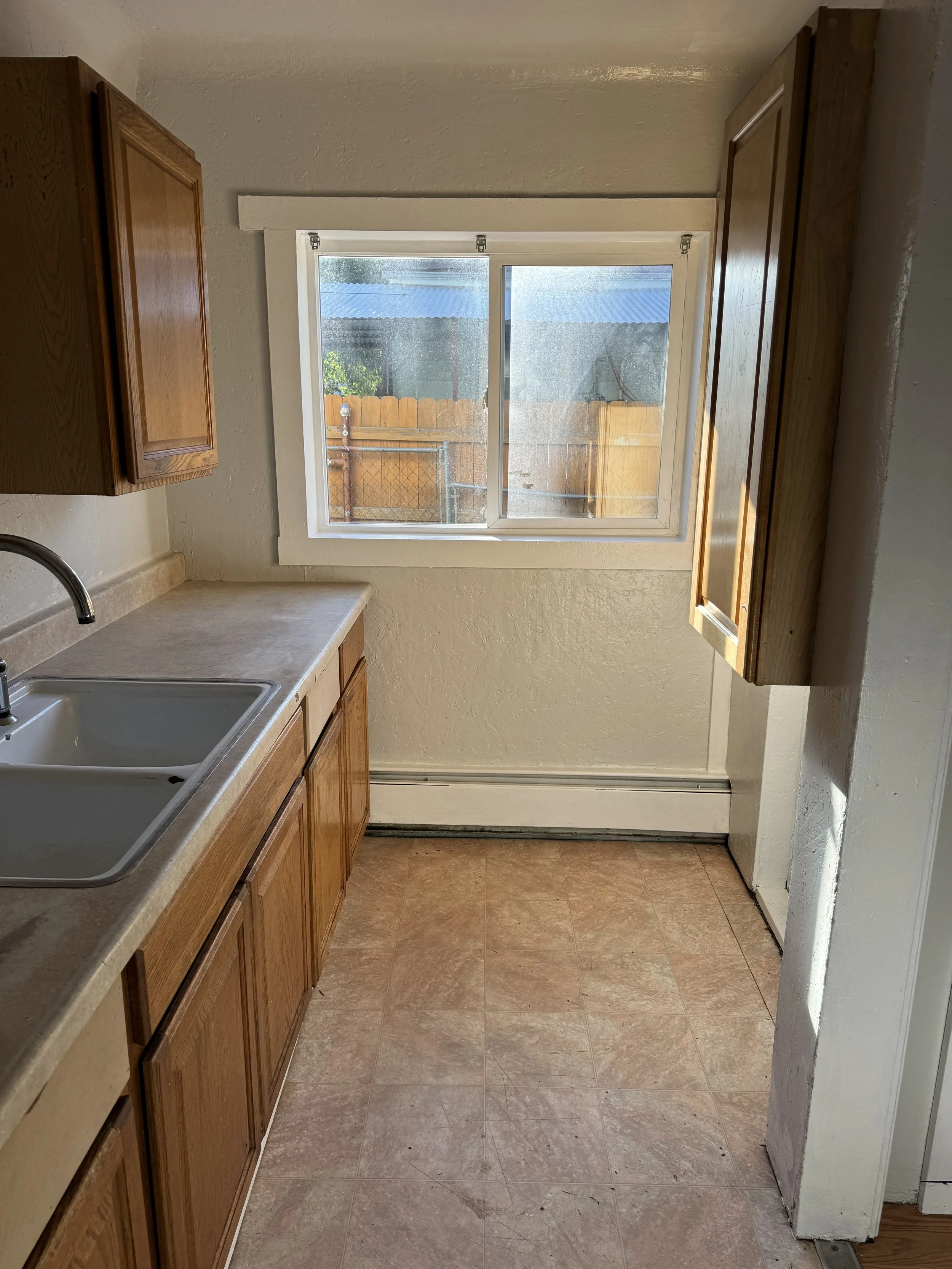 Empty kitchen with wooden cabinets, beige countertop, white sink, and a window looking out to a fenced backyard.