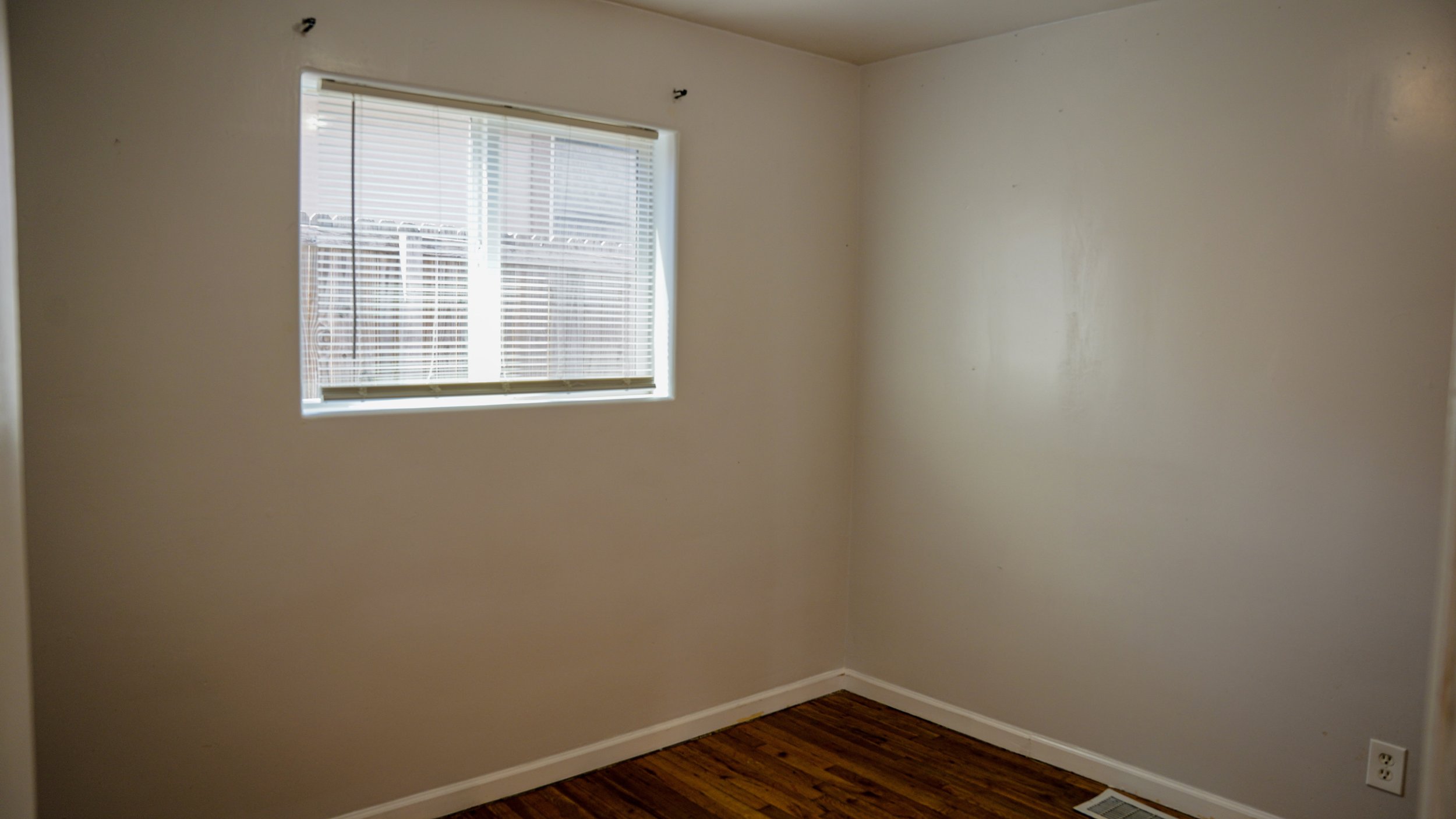 Empty room with a window with blinds, wood flooring, and a power outlet near the baseboard.