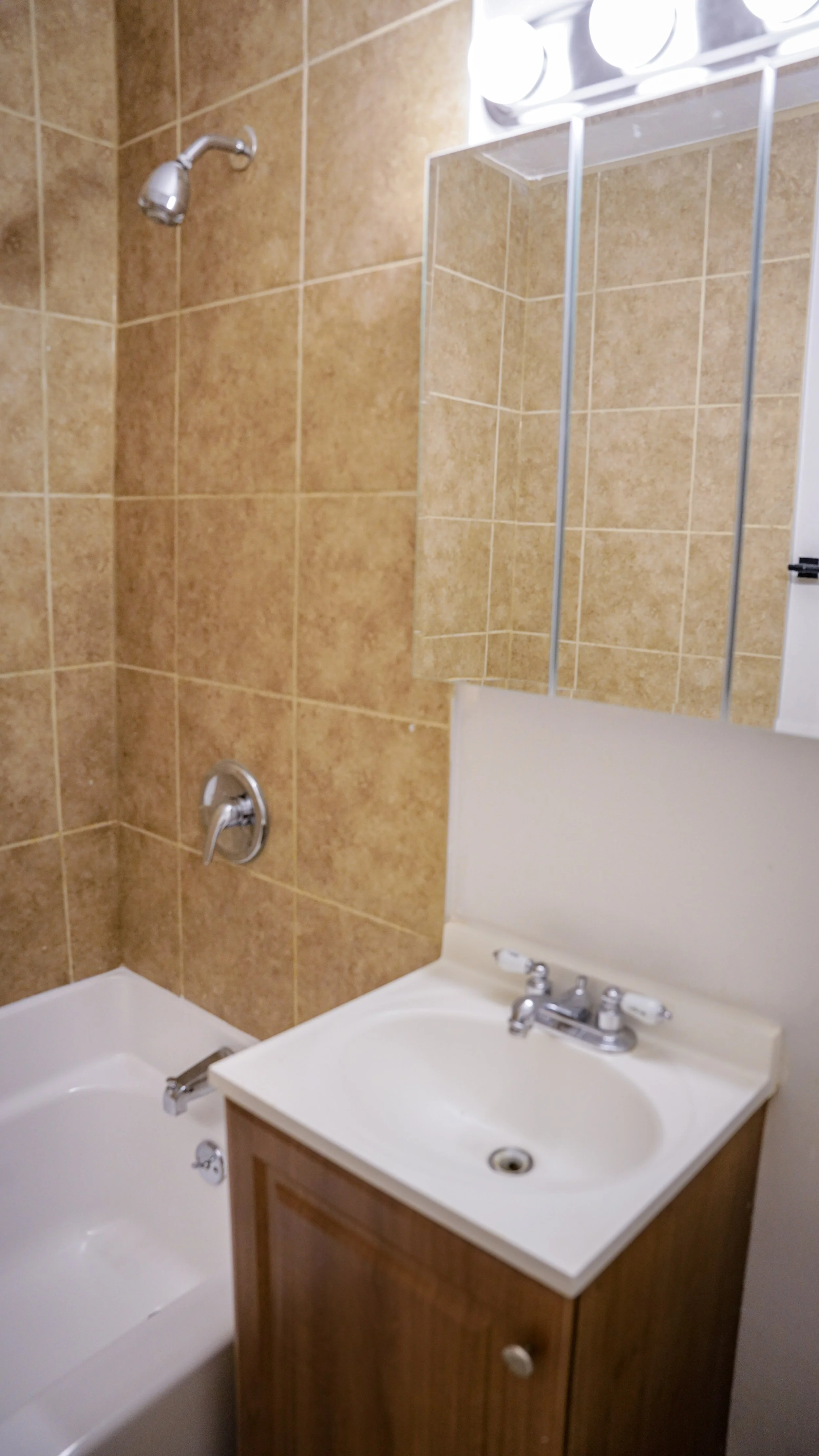 Small bathroom with a bathtub, shower head and faucet, a white sink with a wooden cabinet, a mirrored medicine cabinet, and beige tiled walls.