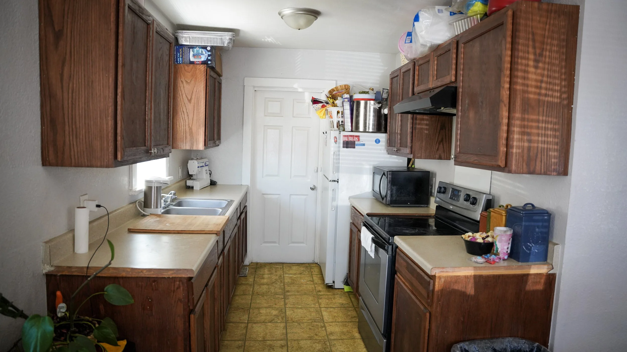 A kitchen with wooden cabinets, a white countertop, a stainless steel sink, a microwave, a stove, and various small appliances and items on the counters. There is a white door at the back and a small plant in the lower left corner.