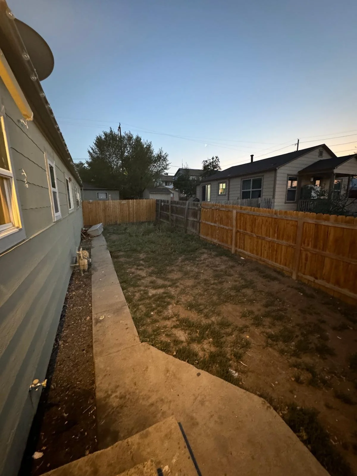 Backyard with a concrete pathway on the left, a grassy area with patches of dirt, and a wooden fence around the yard. There are houses visible in the background during dusk.